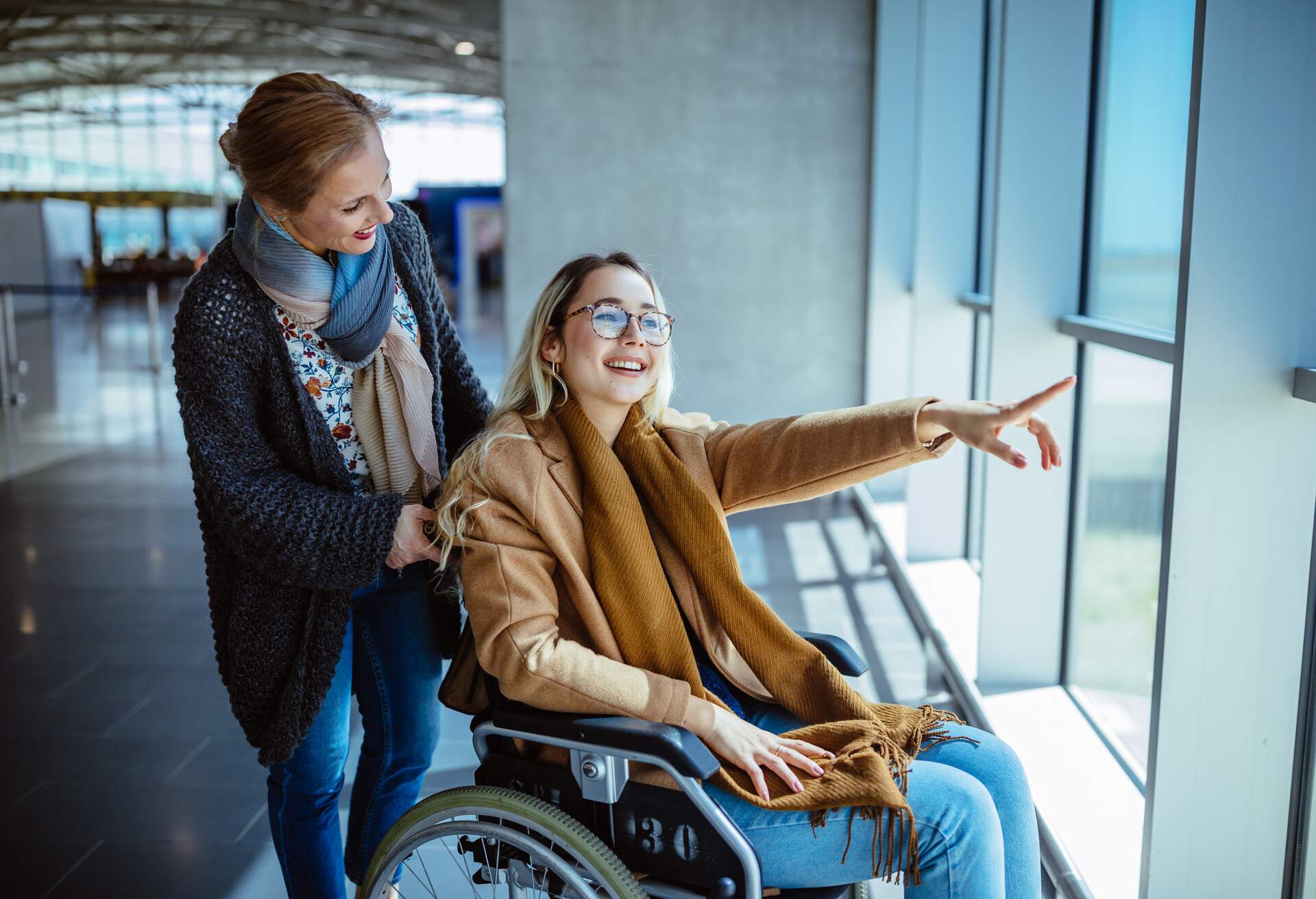 A woman pushes a young woman sitting in a wheelchair inside an airport terminal.