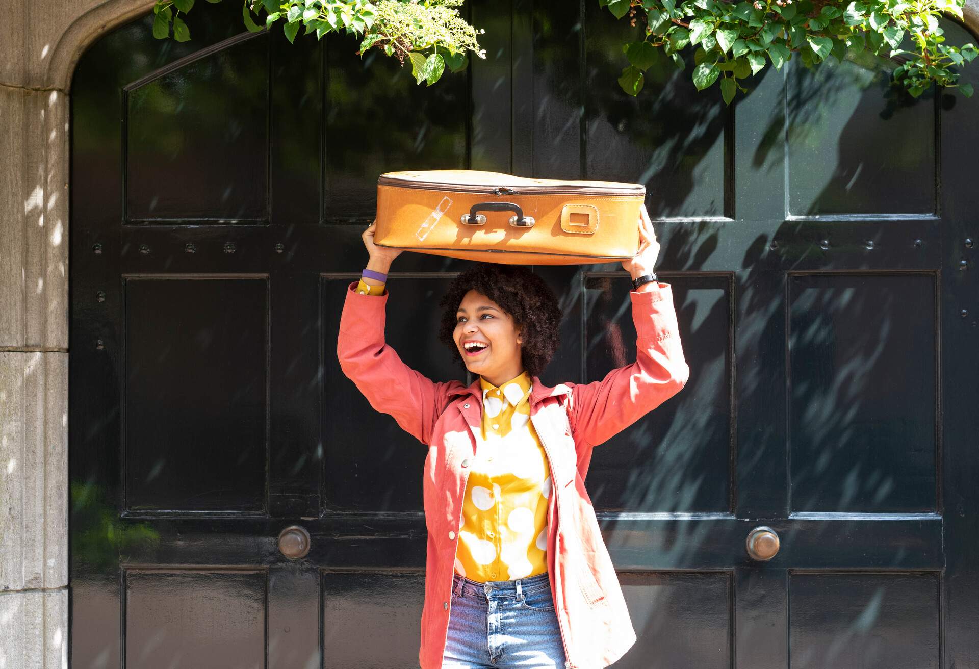 A young woman standing behind a black door, smiling as she lifts her suitcase over her head.