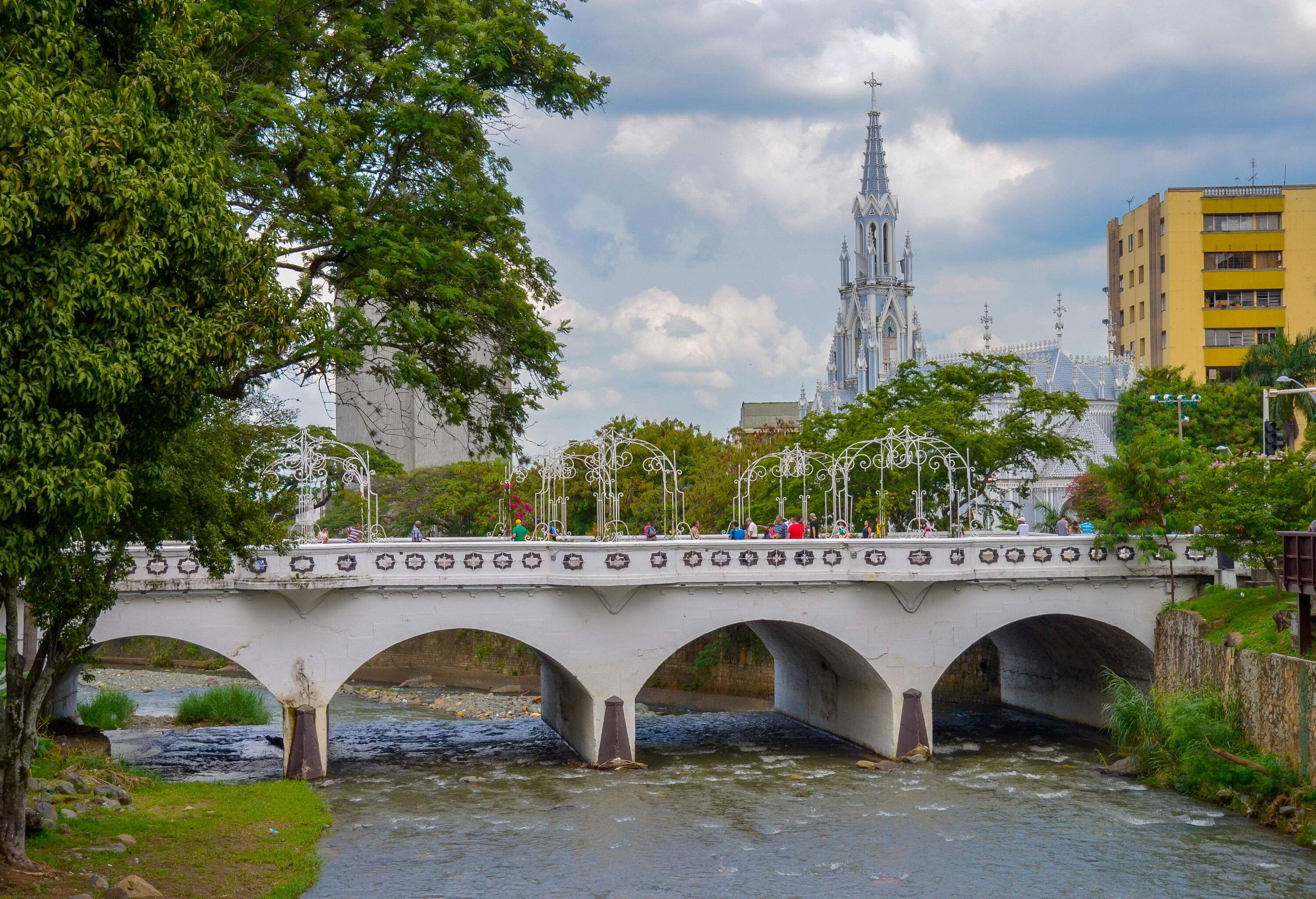 Pedestrians cross a white arch bridge across a calm river with views of a towering church steeple in the background.