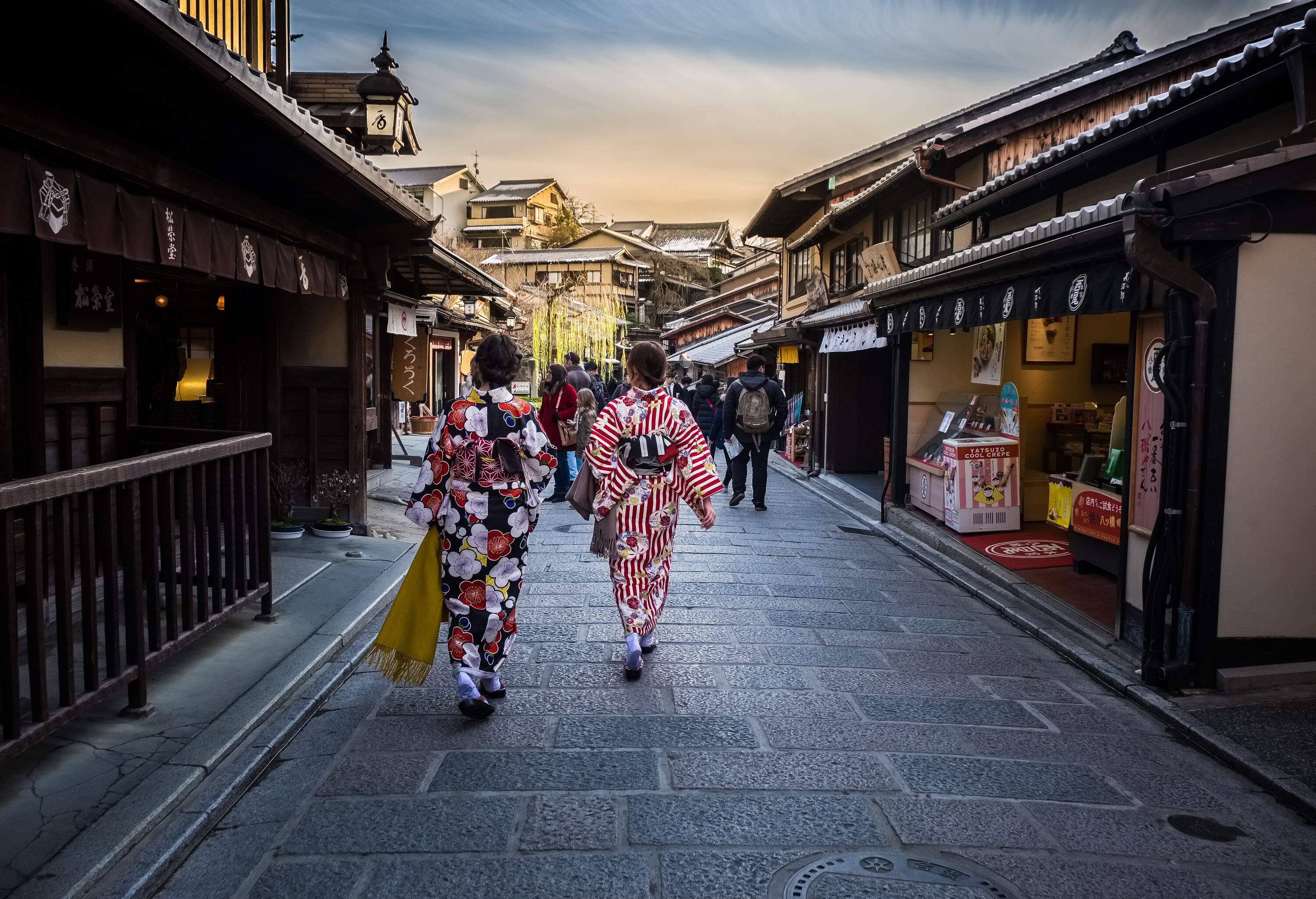 Two ladies in colourful floral kimonos walking in a busy street in the middle of houses.