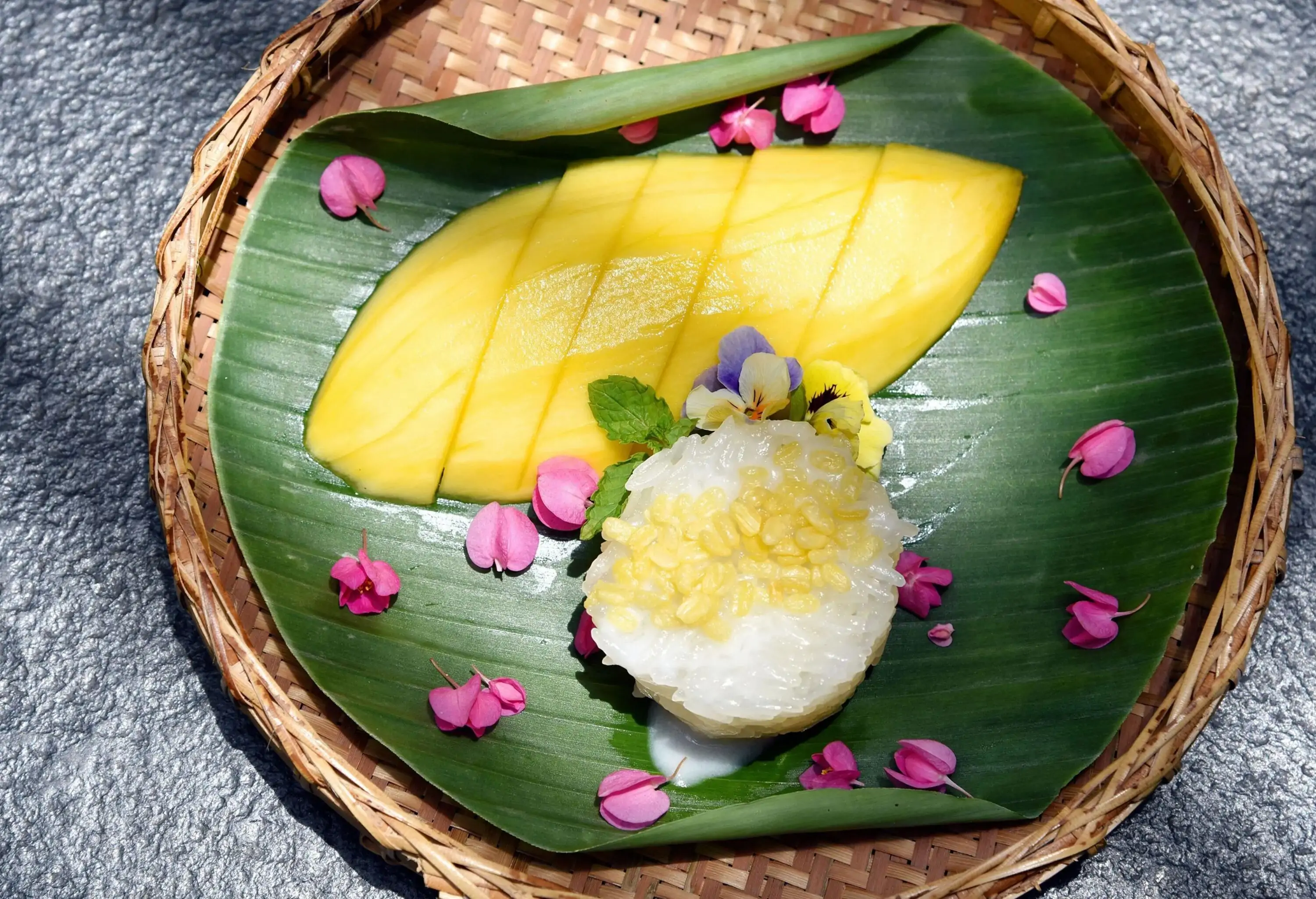 A delightful dessert of mango and sticky rice is served on a bed of fragrant banana leaves, accompanied by a rich and creamy coconut milk sauce and crunchy beans, all elegantly presented on a traditional winnowing tray.
