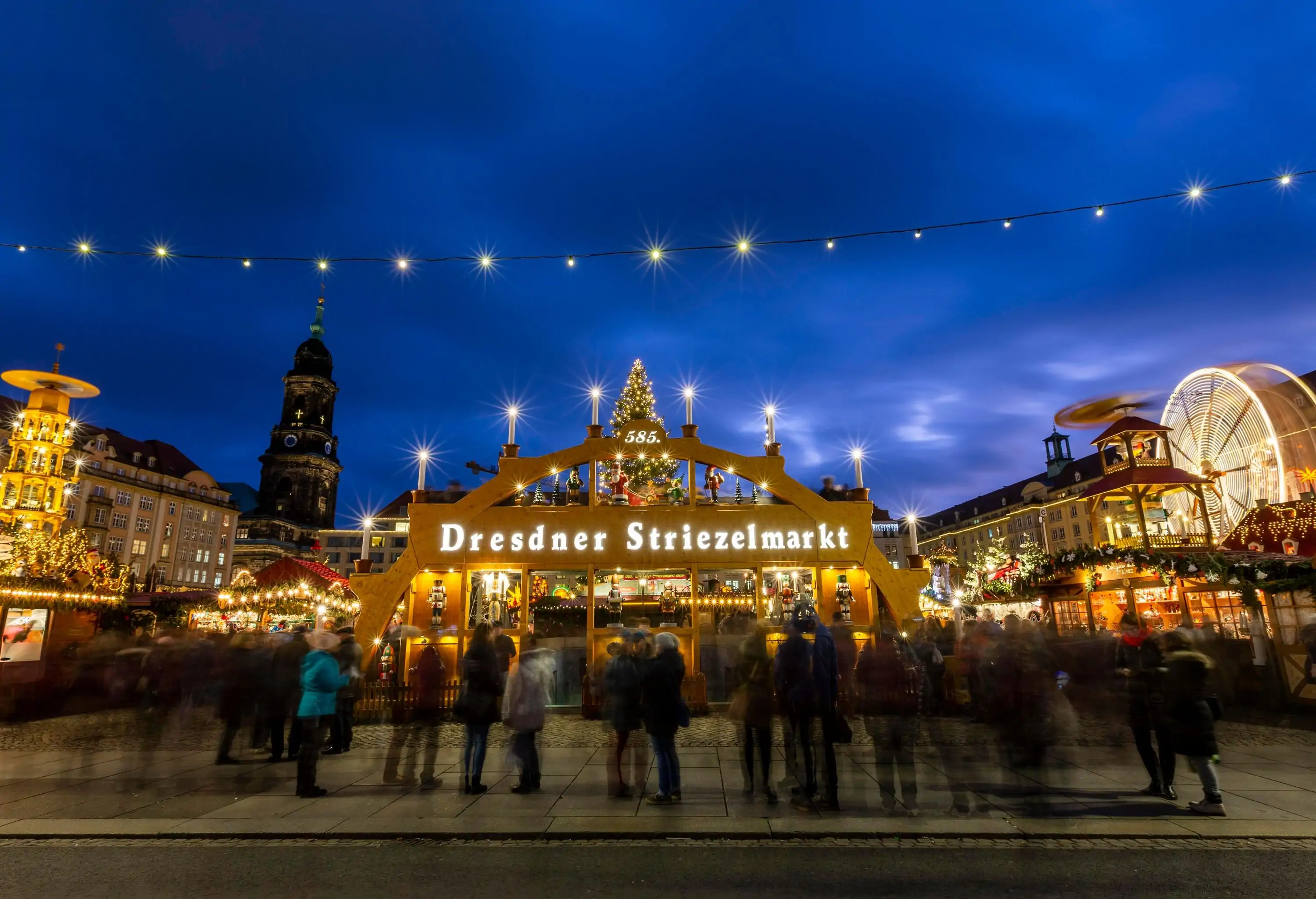 An illuminated golden arch gateway of a Christmas market in a square enclosed by tall buildings in Dresden.