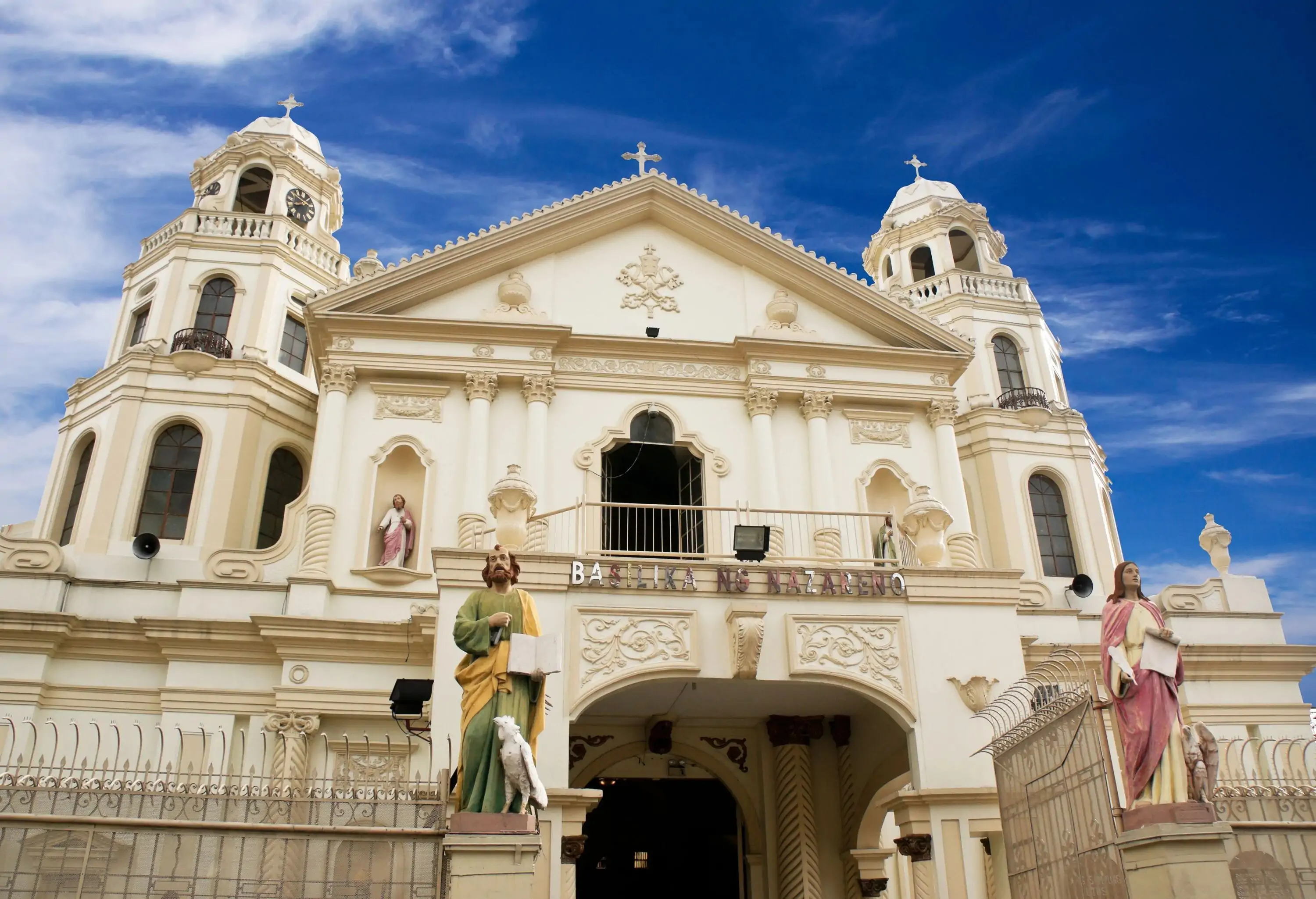 Quiapo Church facade, a stunning blend of Spanish Baroque and Neo-Gothic styles, adorned with intricate carvings and towers, is fronted by a fence topped with statues.