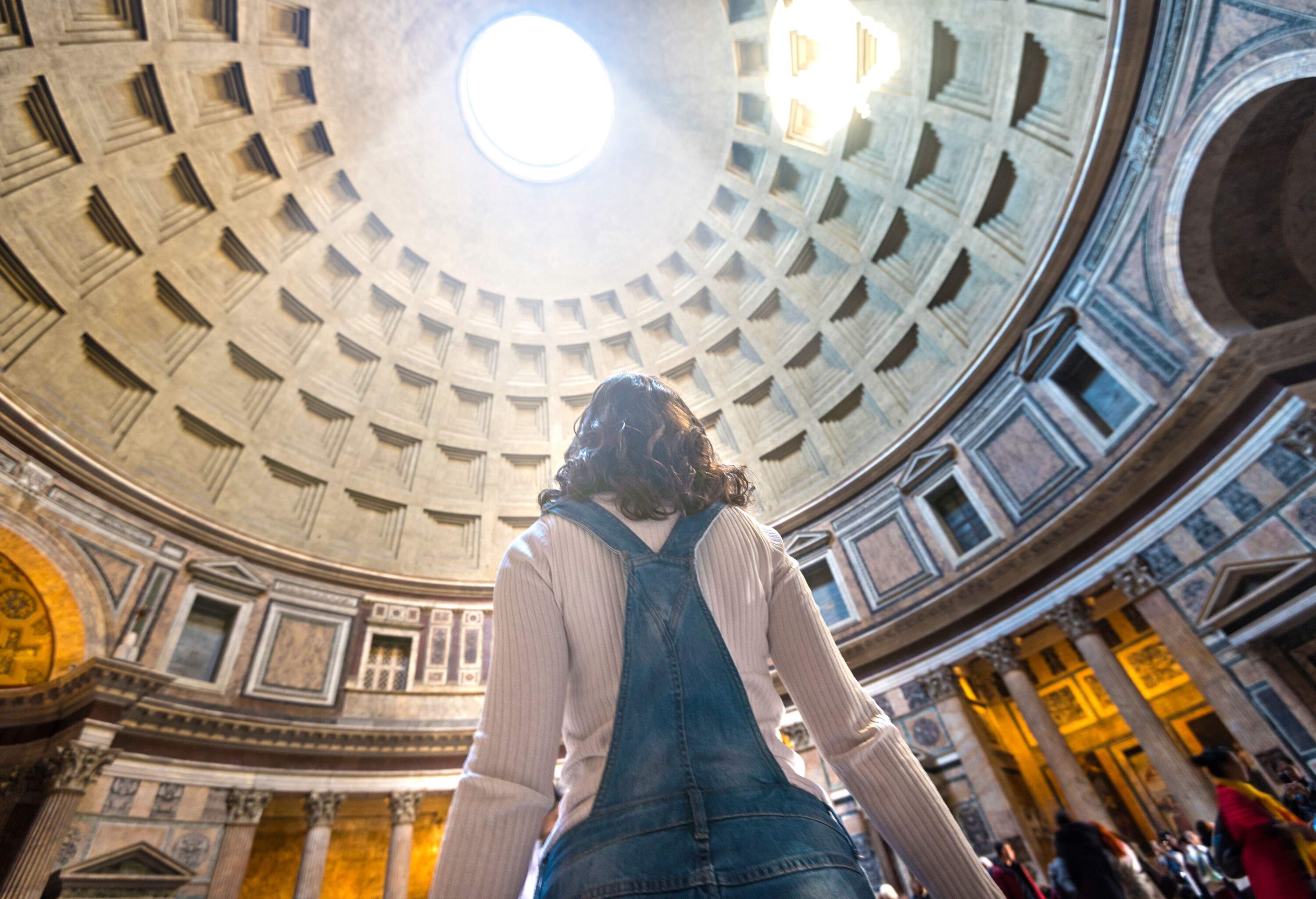 A woman is standing inside a building with a coffered dome with light passing through the central oculus.