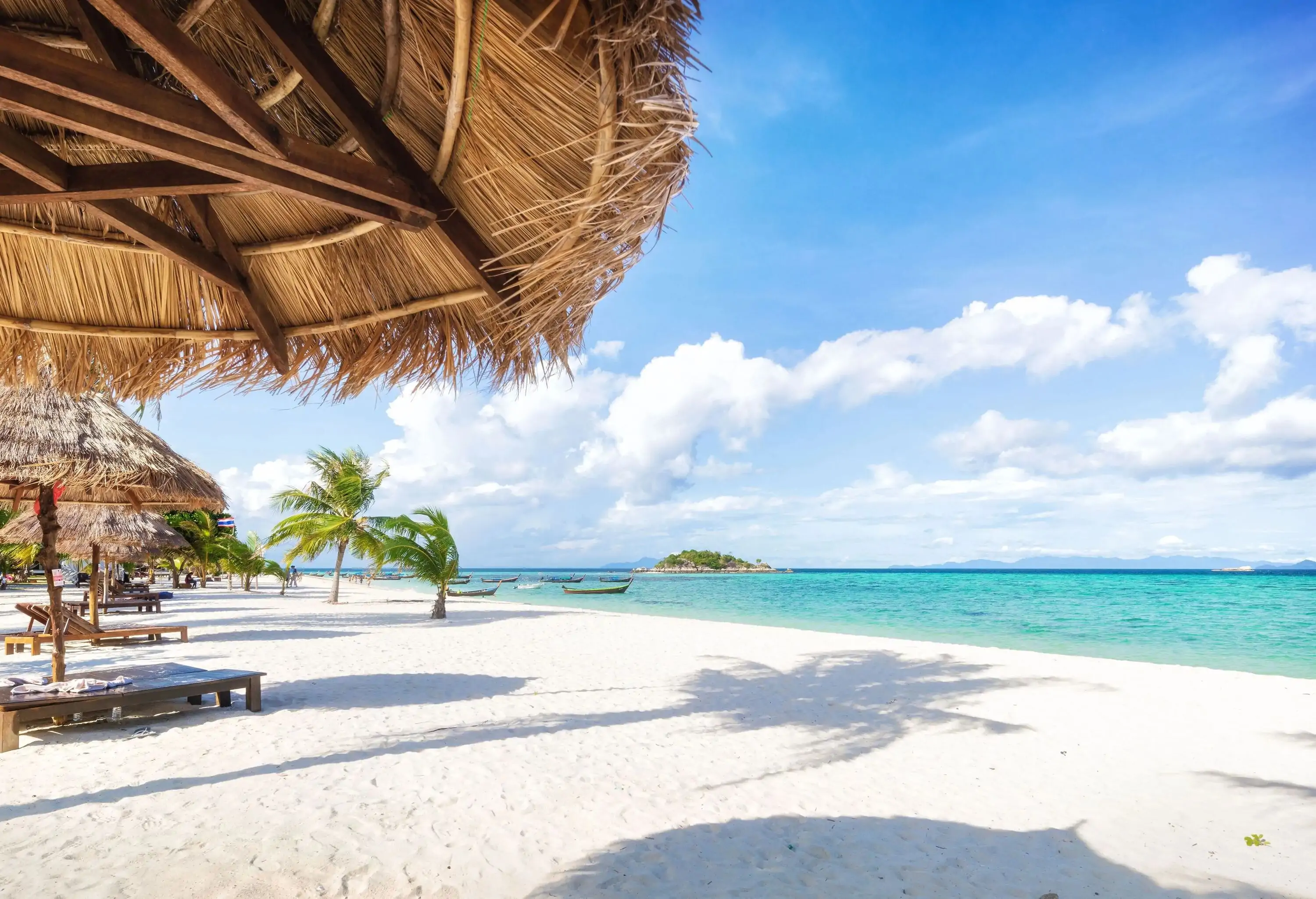 Thatched parasols provide shade on a pristine white-sand beach, with the tranquil sea lapping gently at the shore.