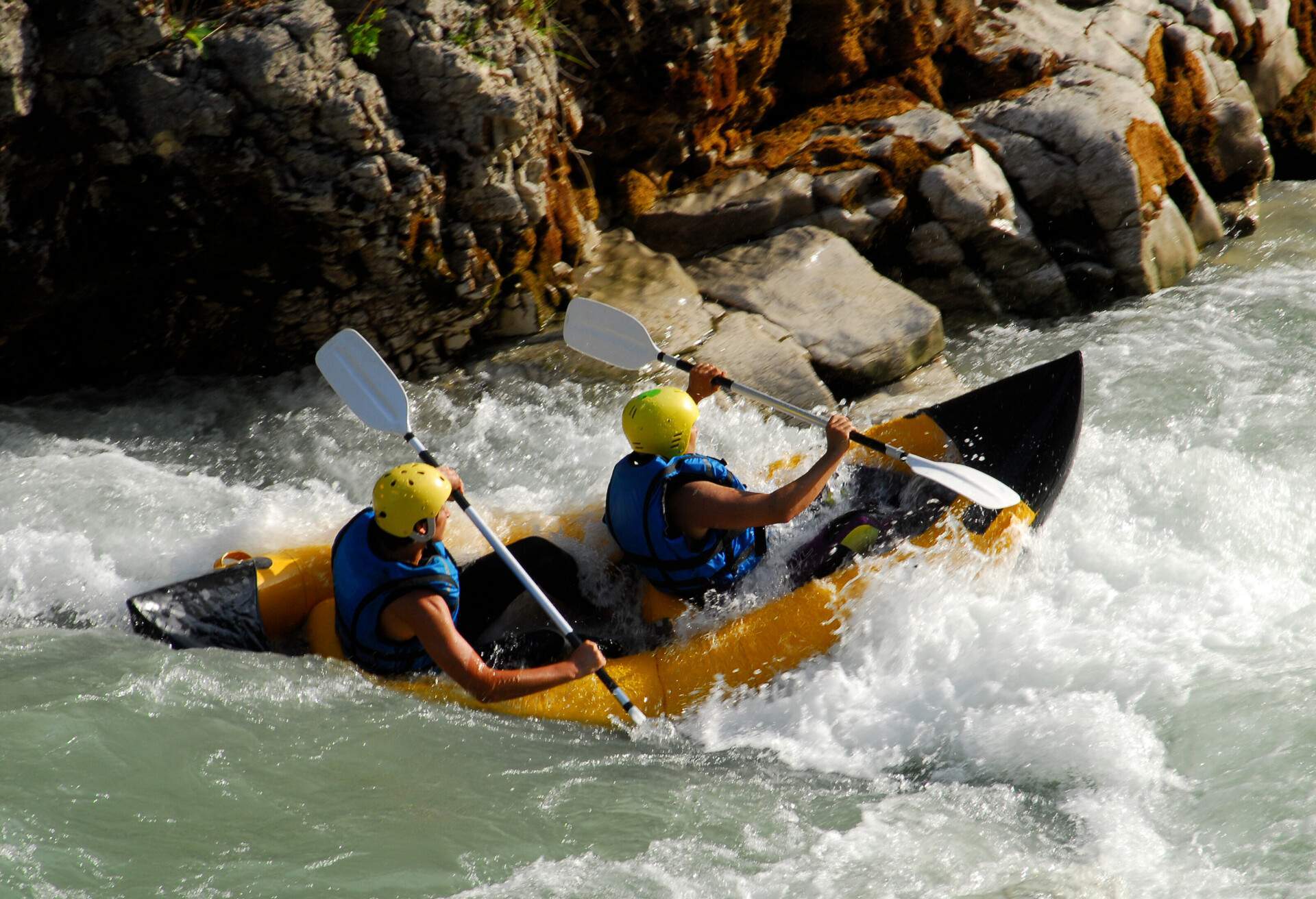 Two people in a yellow boat with life jackets and helmets are rafting on a fast river.