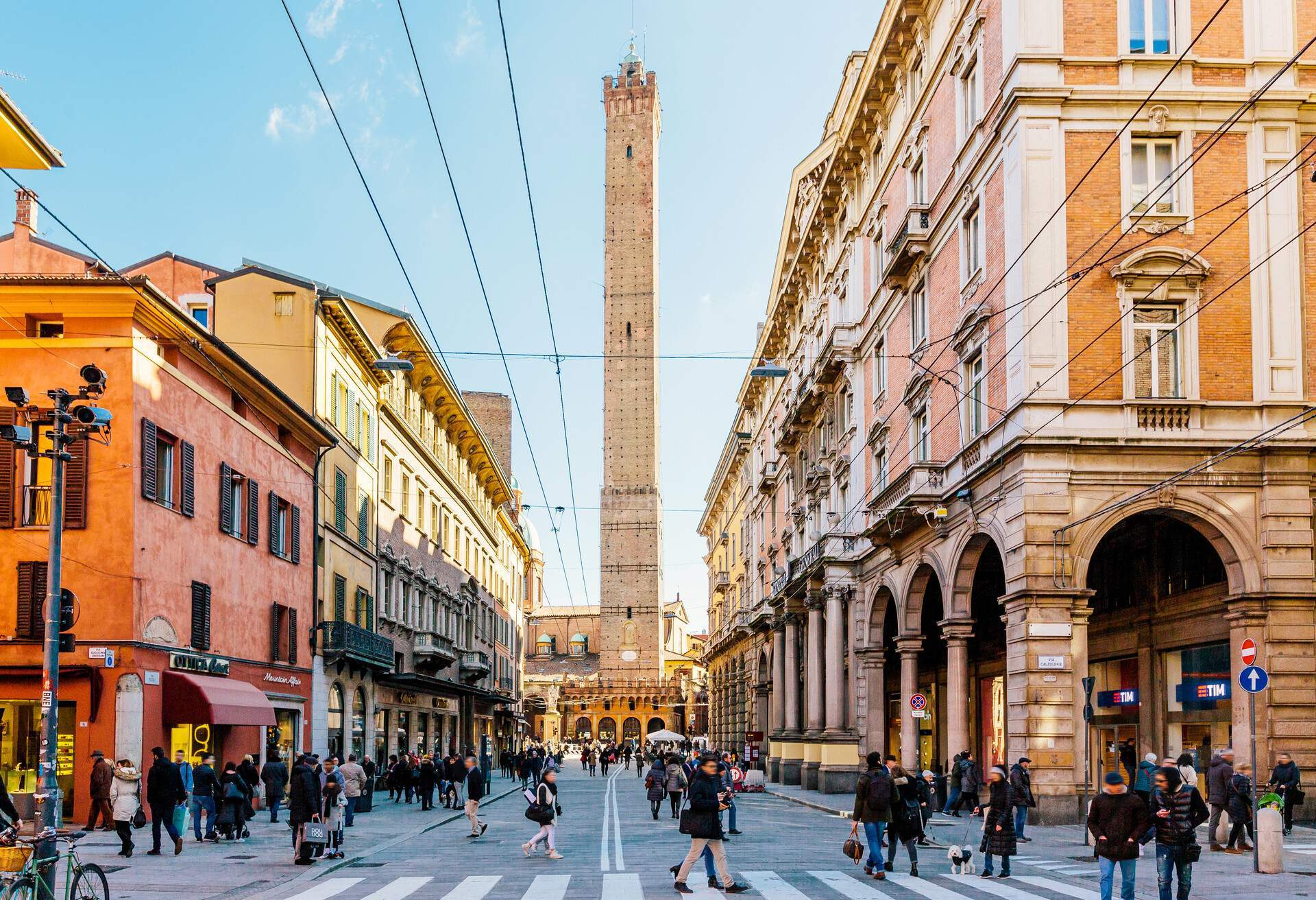 A bustling street lined with colourful and classic commercial buildings with a towering medieval structure in the centre.