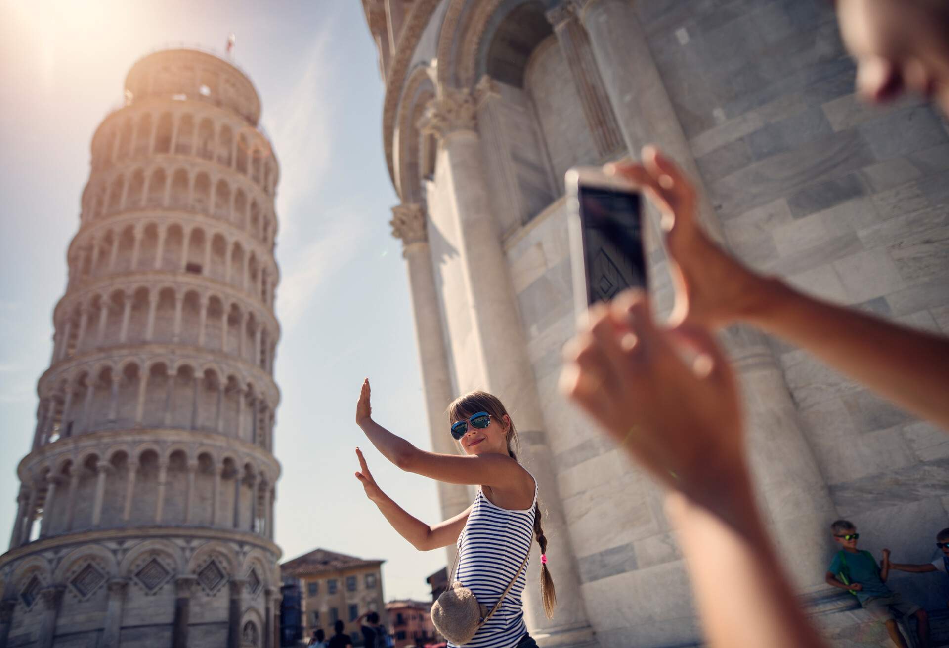 A phone in hand snapping pictures of a young woman posing with her arms raised near the Tower of Pisa.