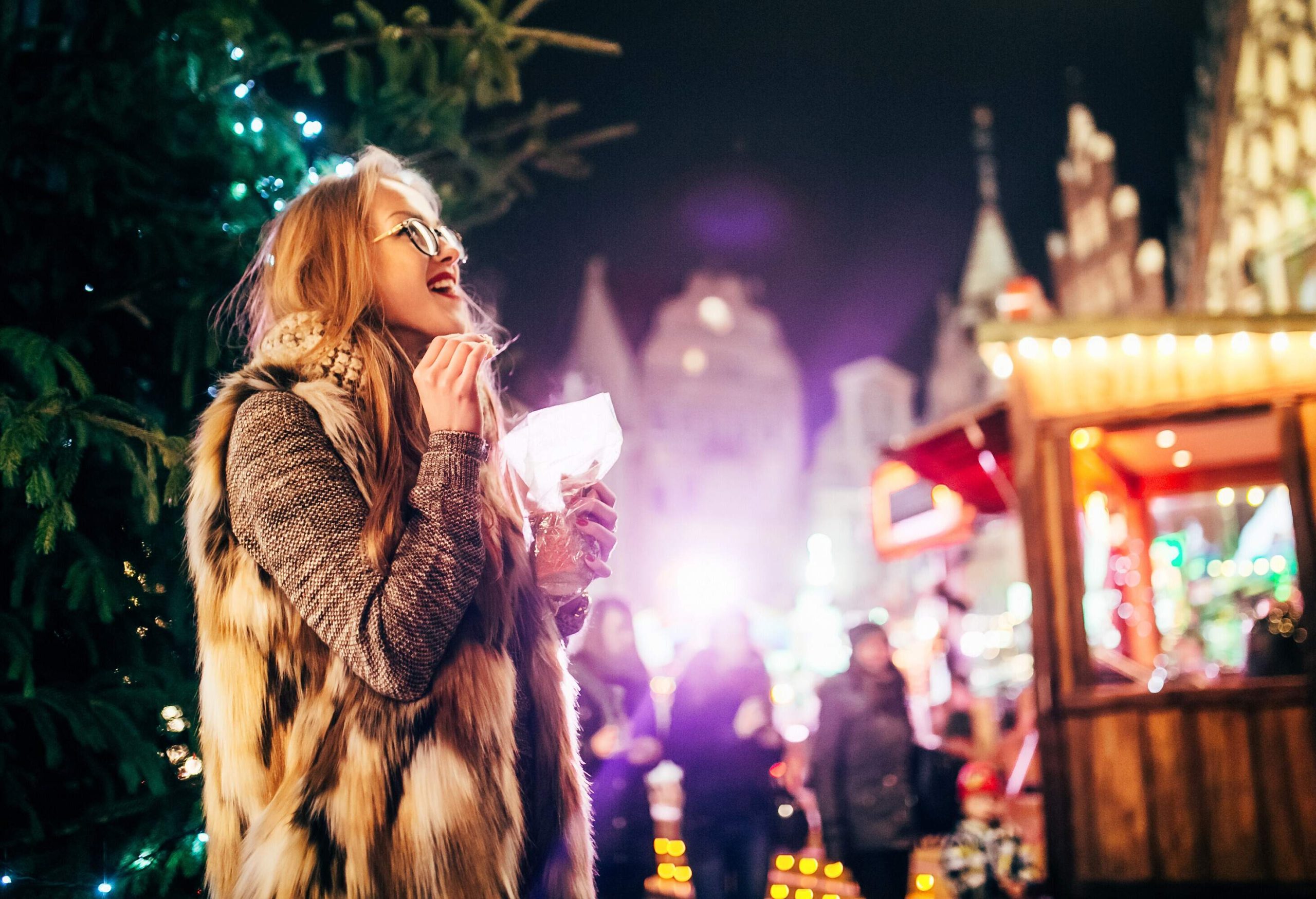 A blonde woman in fur and glasses smiles as she looks up at holiday lights while holding a snack.