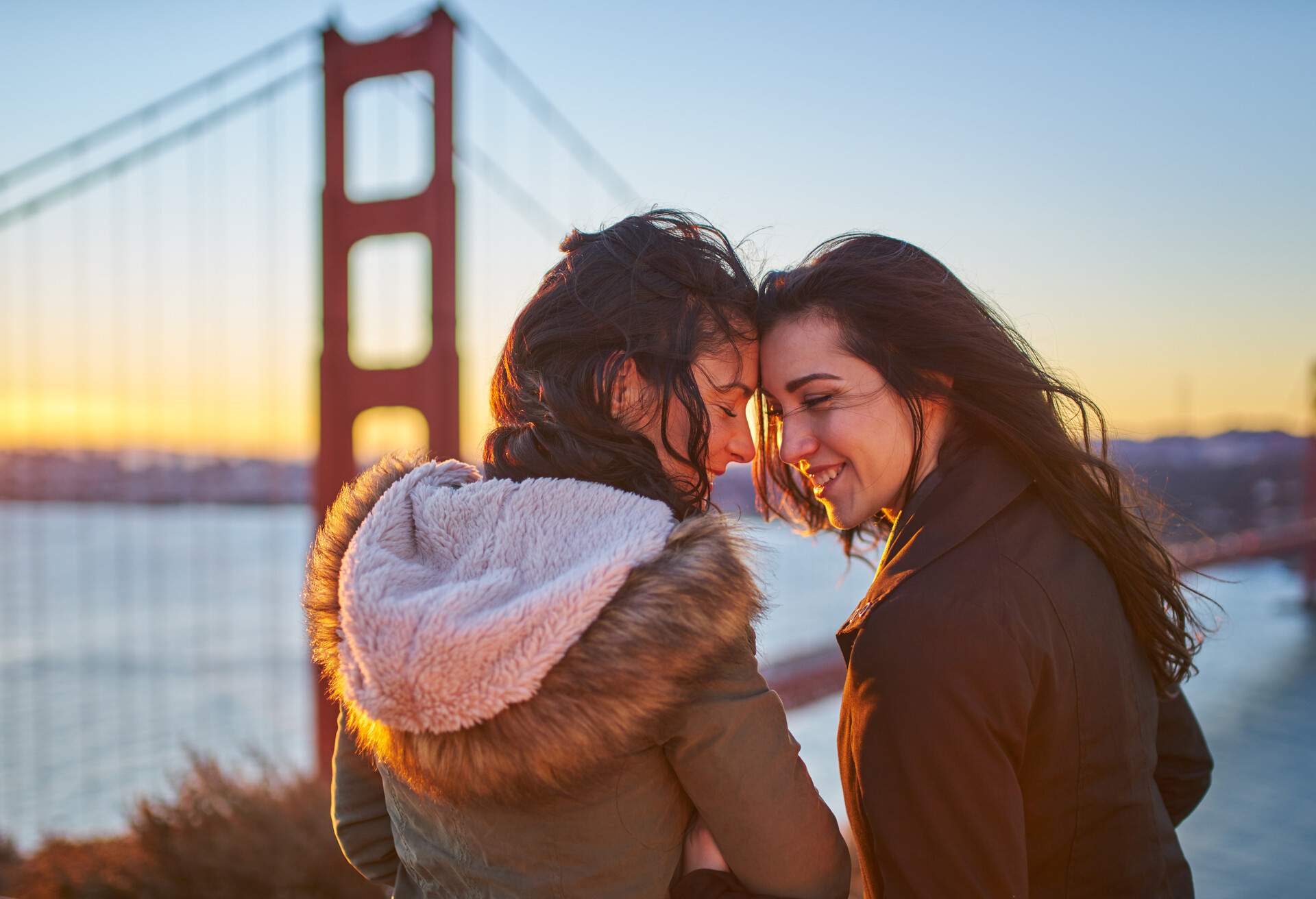 A flirty couple in love share an intimate moment, framed by the spectacular view of the Golden Gate Bridge, as a vivid and colourful sunrise bathes the scene in warm, romantic light.