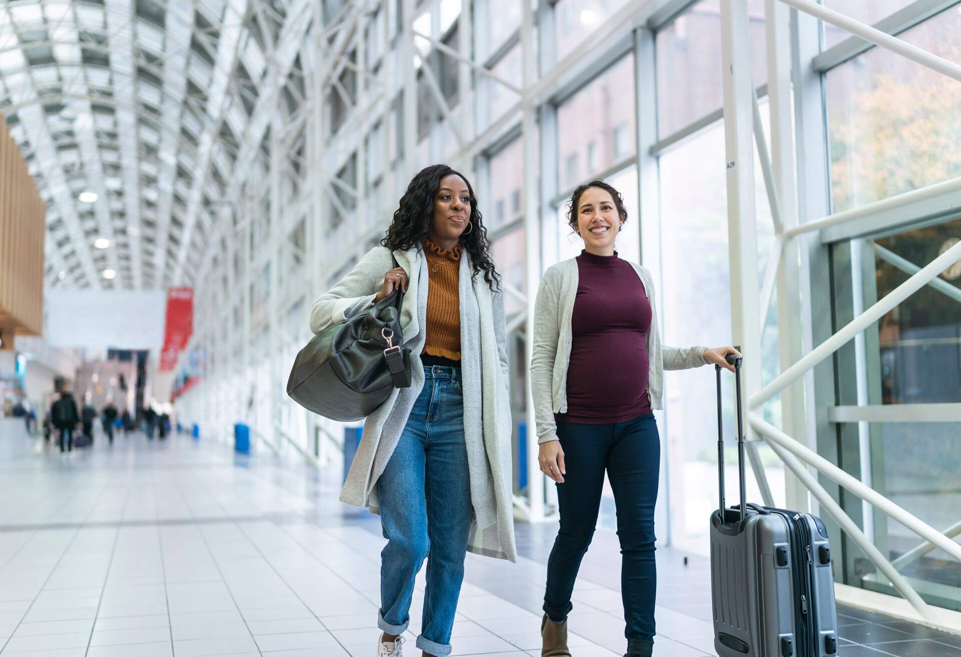 Two female friends are traveling together. One of the women is black and the other is Eurasian. The Eurasian woman is pregnant. The smiling women are talking happily. They are carrying luggage while walking side by side through the airport. Babymoon and safety while traveling pregnant concept.