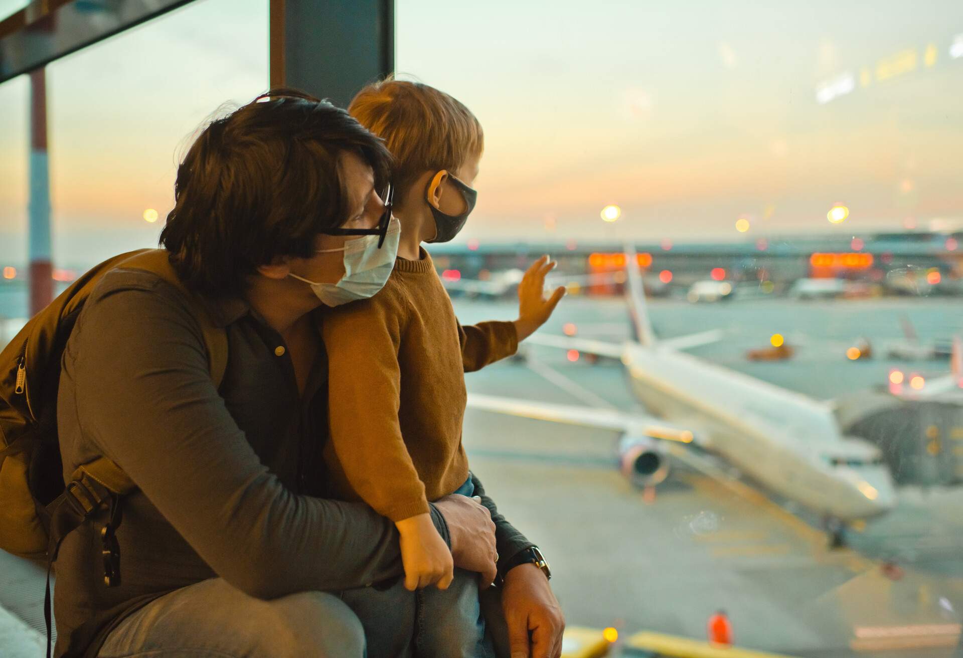 A father and son wearing facemasks in an airport terminal looking at the aeroplanes at the runway.