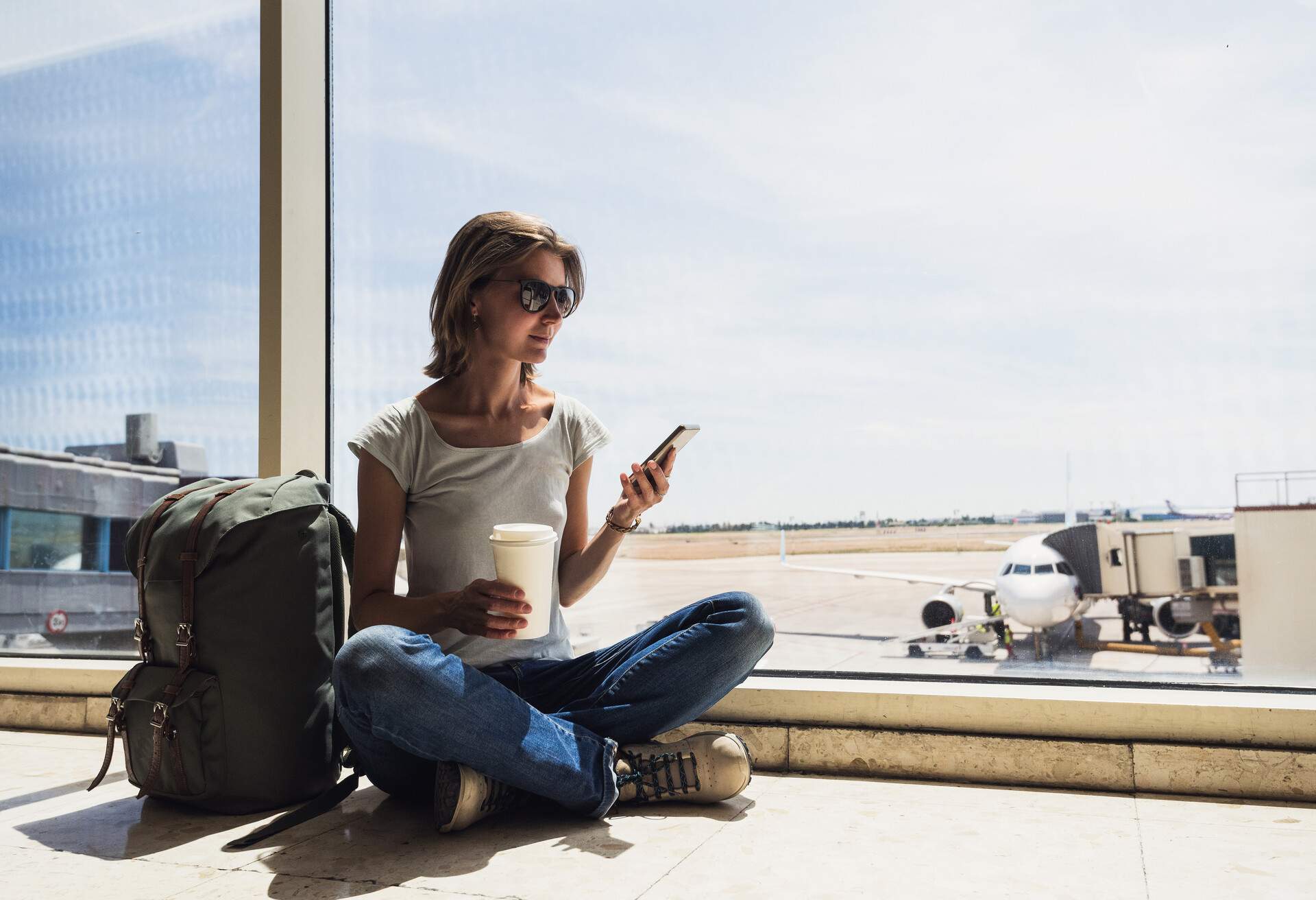 A young girl eagerly awaiting her plane at the airport, comfortably seated on the floor, holding a drink and her phone in her hands, framed by the transparent glass wall of the boarding area.