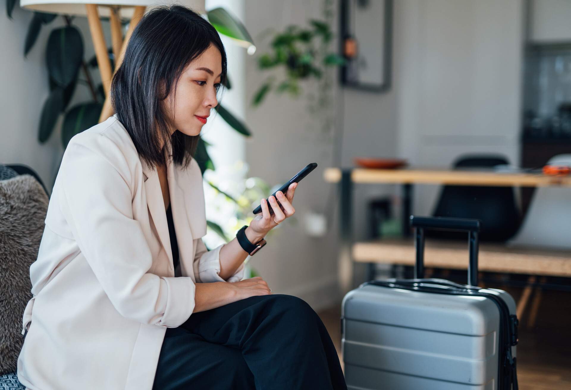 A lady traveller sitting on the sofa near her luggage while using her phone.