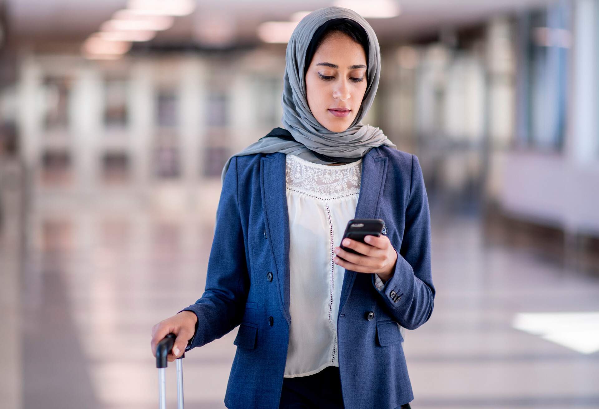 A female wearing hijab looks at her mobile phone while holding a suitcase.