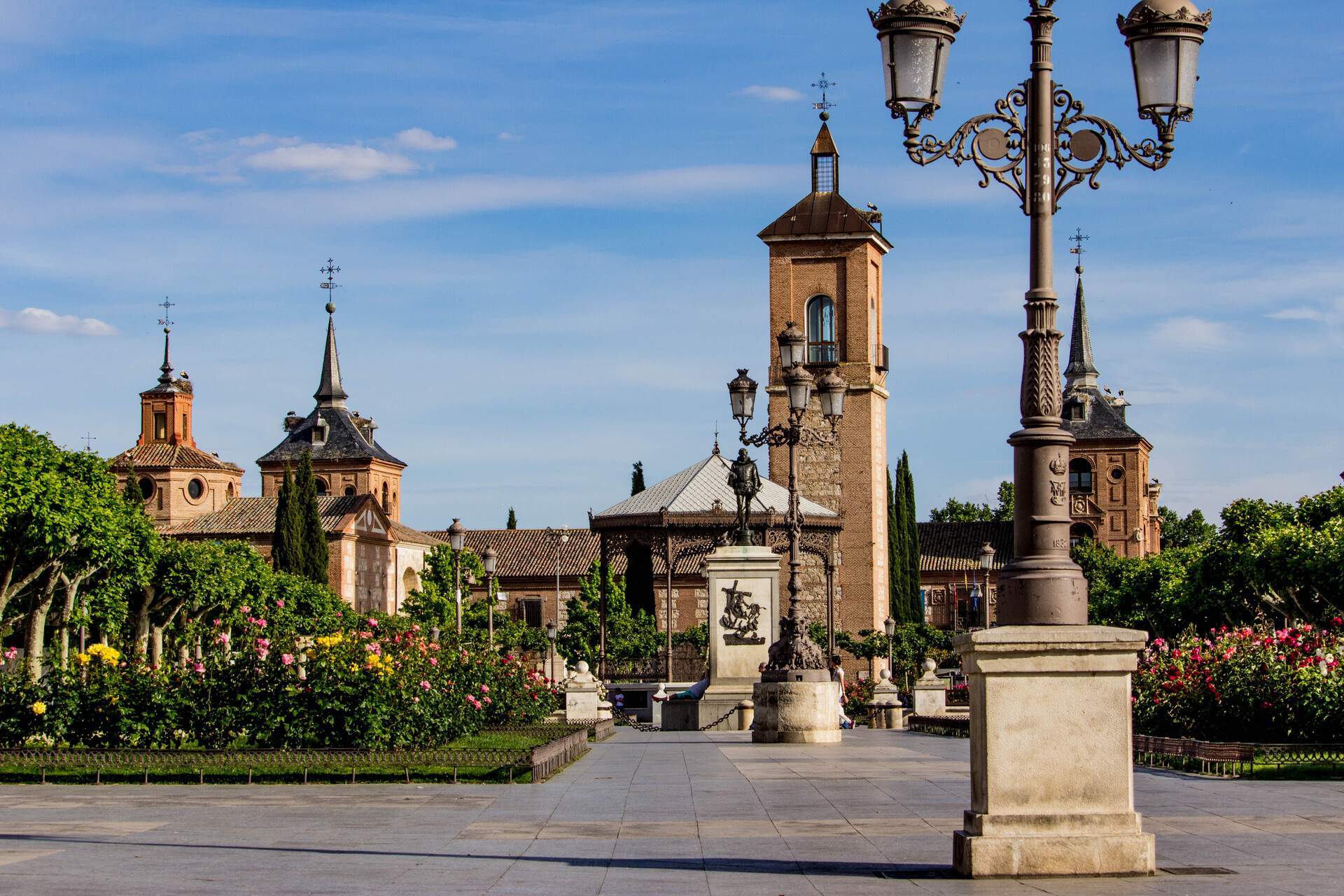 A plaza with a gazebo, lamp posts, and a landscape of colourful flowers and trees with a view of towers with spires.