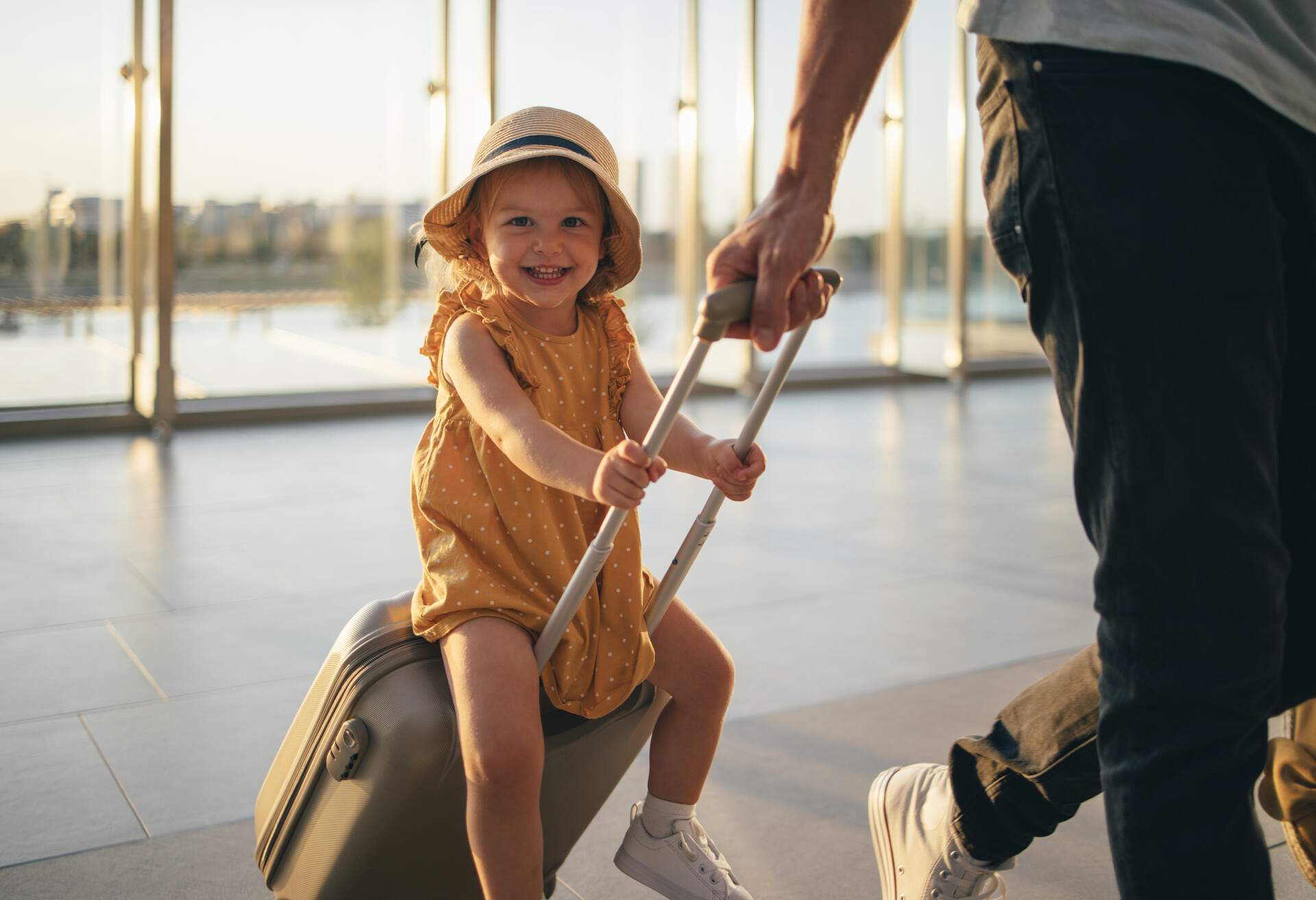 Anonymous father walking with his little daughter sitting on luggage and looking at camera at the airport.