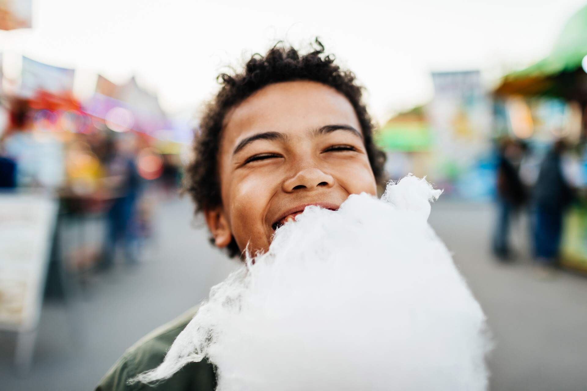 A curly-haired boy smiles as he enjoys eating a cotton candy.