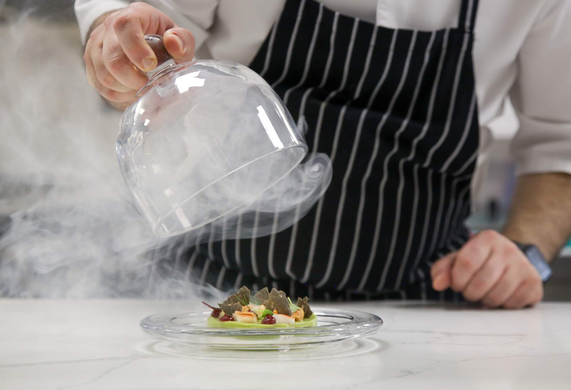 Chef's hand lifts up glass cloche from a plate with hot food and moving smoke at the restaurant.