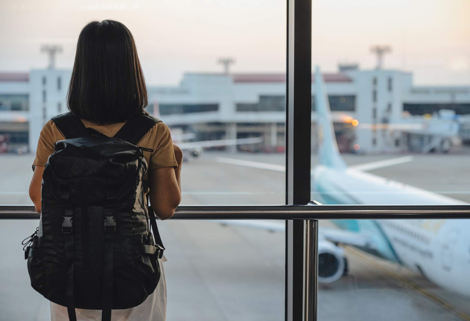 Travel tourist standing with luggage watching sunset at airport window. Woman looking at lounge looking at airplanes while waiting at boarding gate before departure.