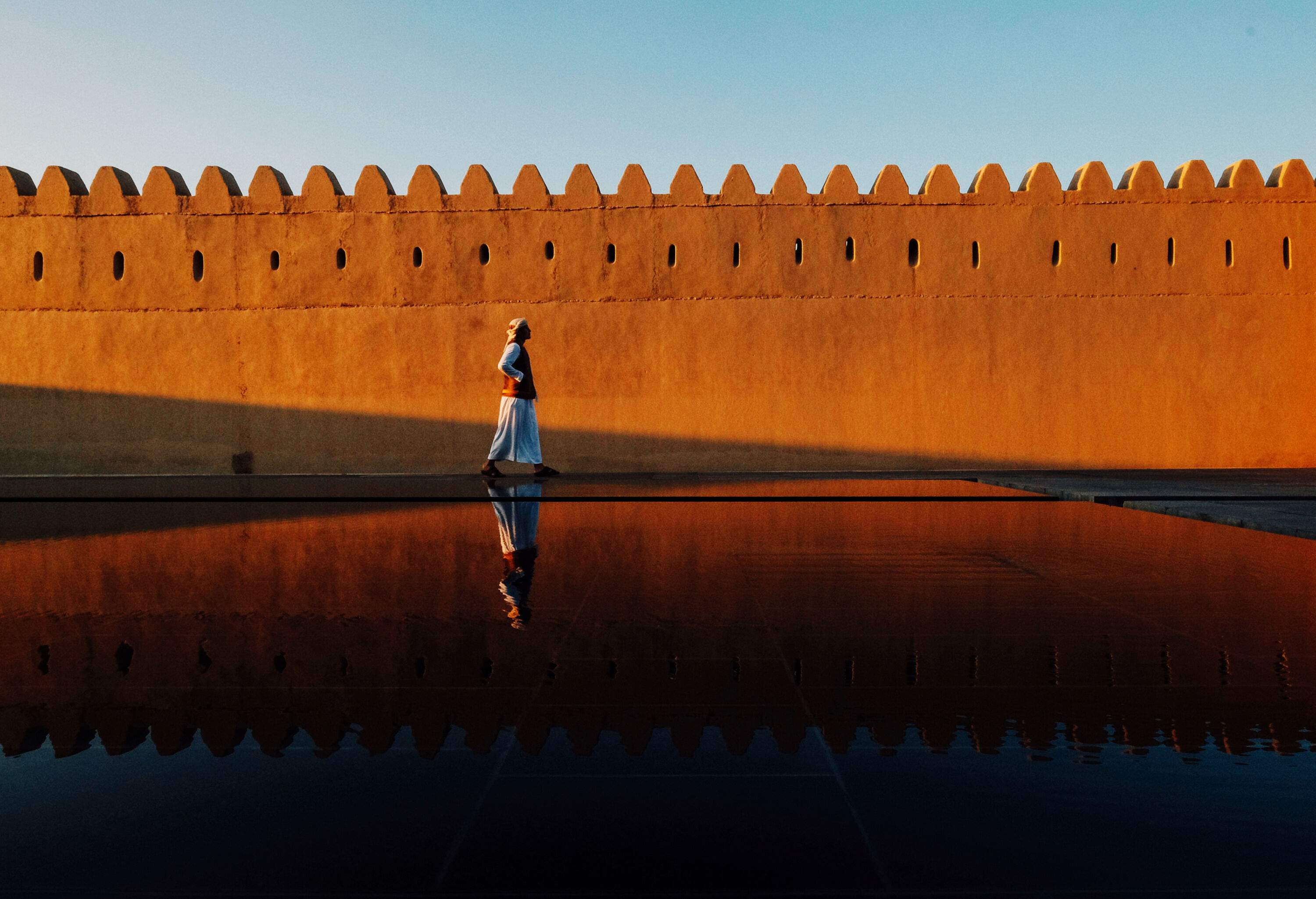 A man walking along a fortress wall with his reflection on the pool of water beside him.