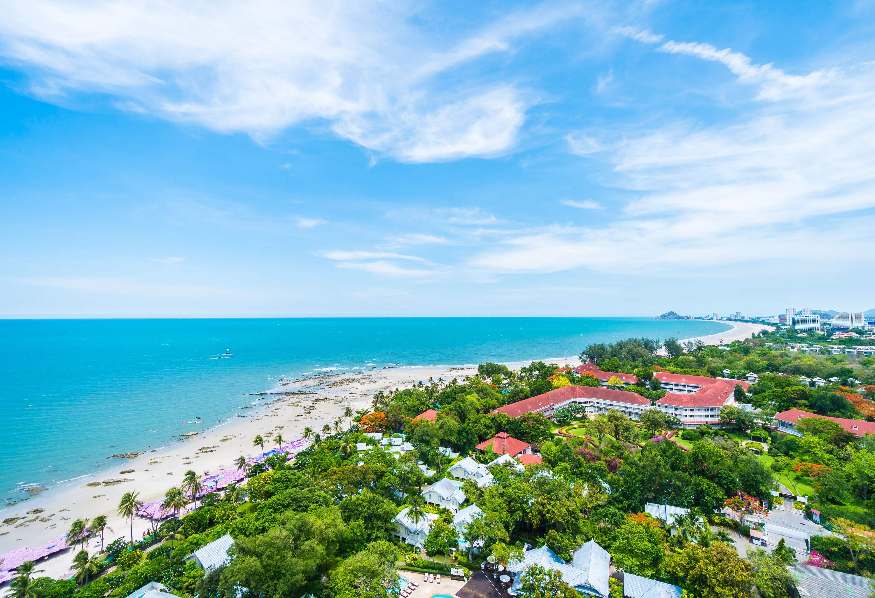A picturesque lush coastal city with a white sand shore by the tranquil turquoise sea against the cloudy blue sky.