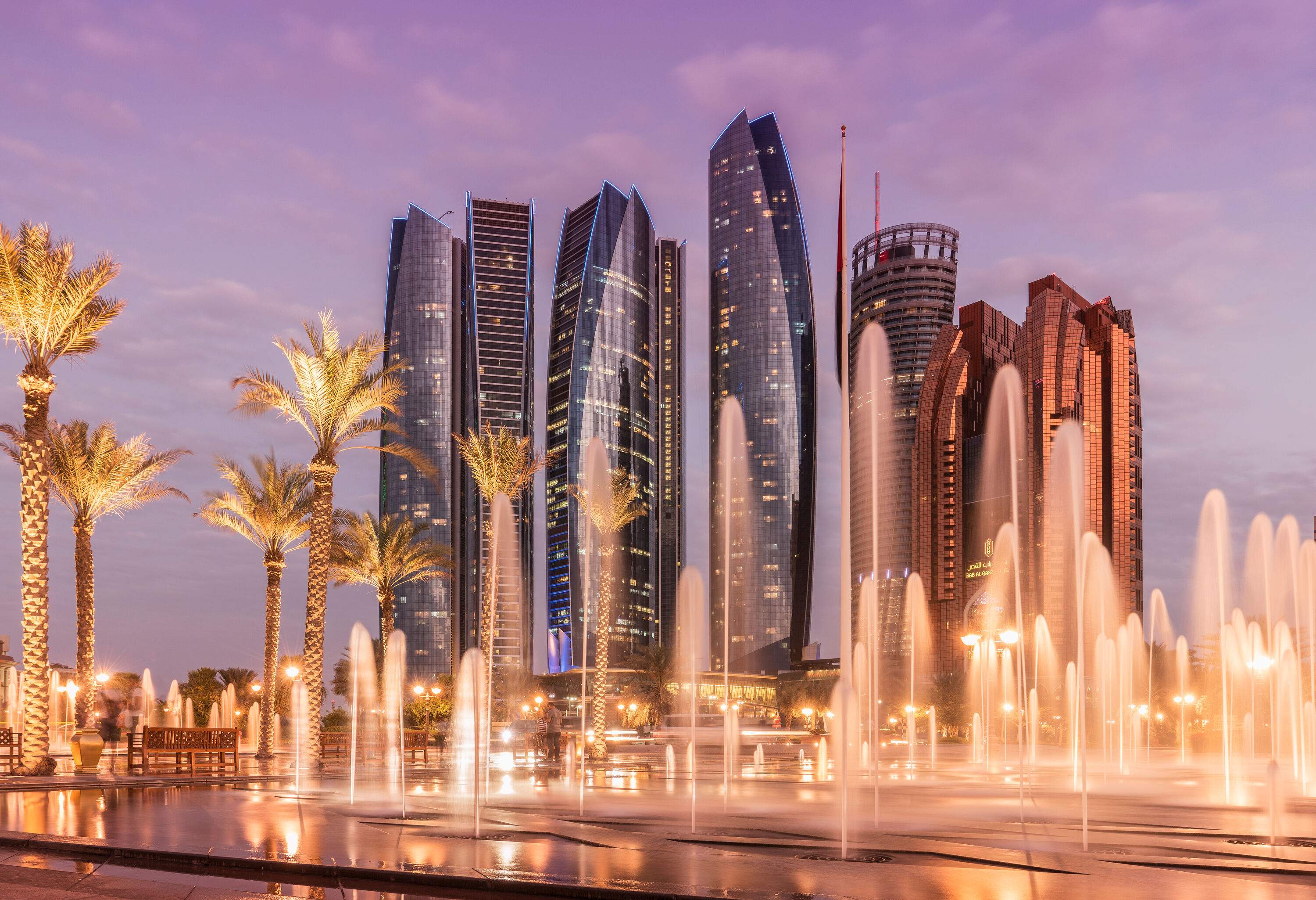A night-time view of spouting fountains beside a grove of illuminated palm trees against a backdrop of city skyscrapers.