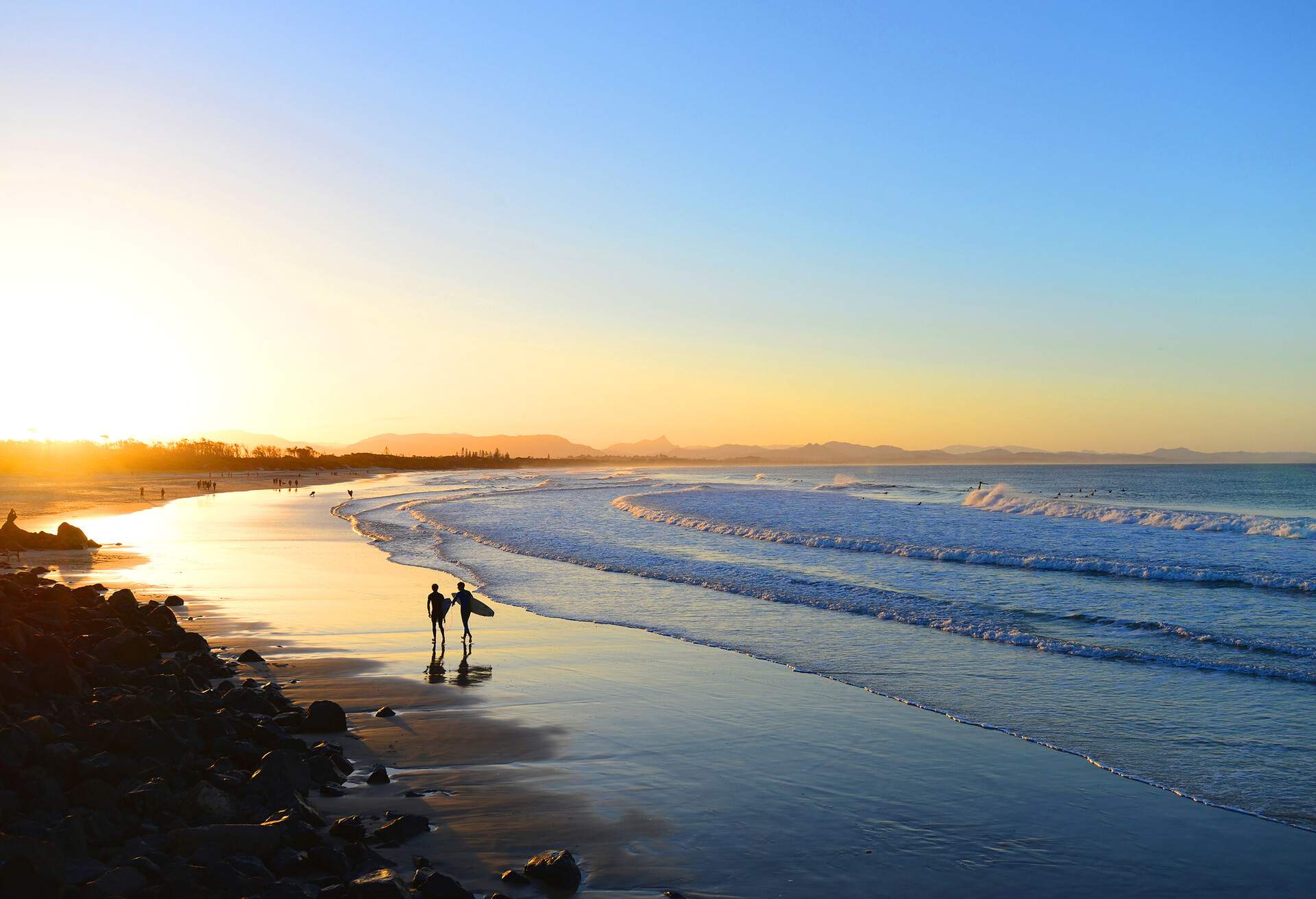 A silhouette of two surfers walking along the beach, with waves gently lapping on the shore, set against the backdrop of a beautiful sunset.