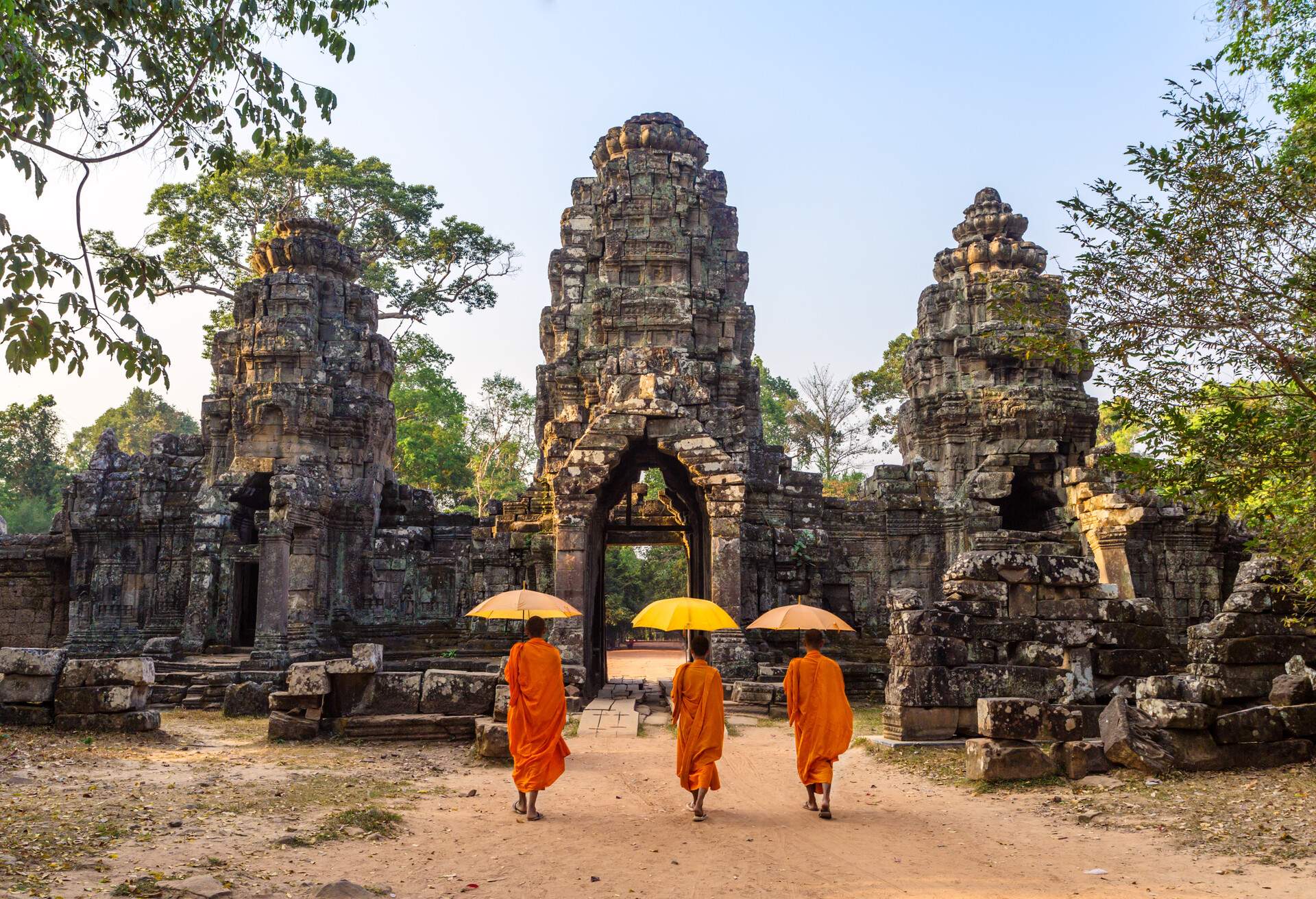 Three Buddhist monks, sheltered by their umbrellas, gracefully traverse the historic stone gate entrance of the majestic Angkor Wat temple complex.