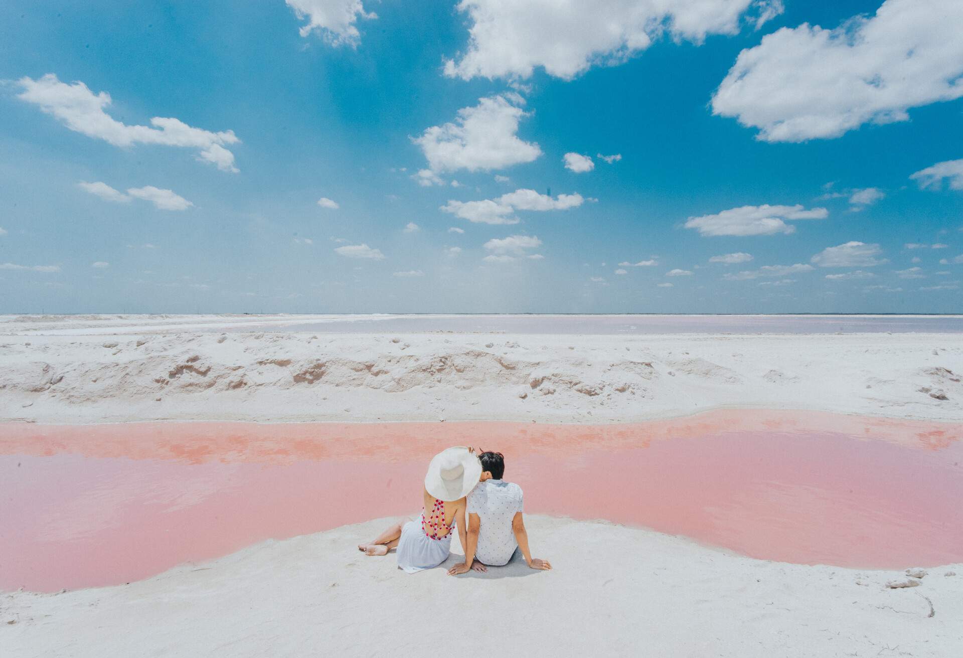 A lovely sweet couple sits on the white bank of a stunning pink lagoon.