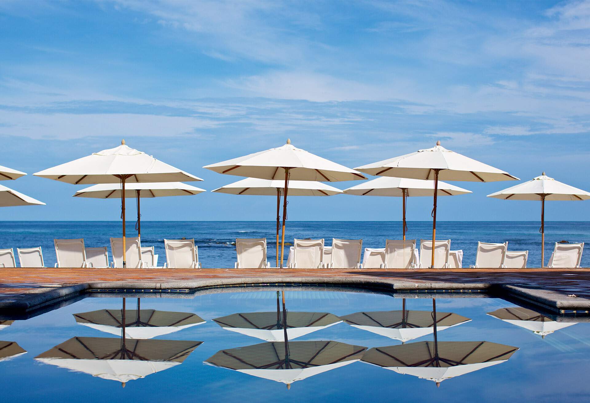 White umbrellas with chairs reflected on the pool's water surface facing the sea.