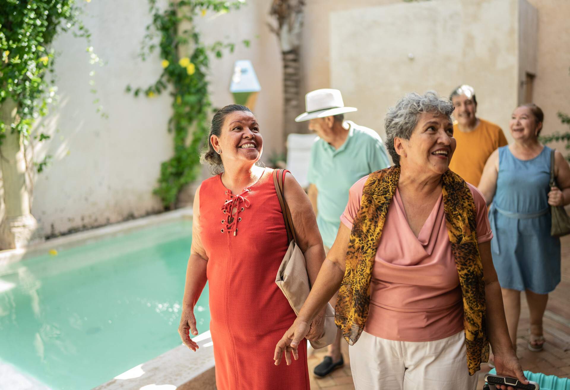 A group of senior friends strolls by a shimmering pool as they arrive at their accommodation.