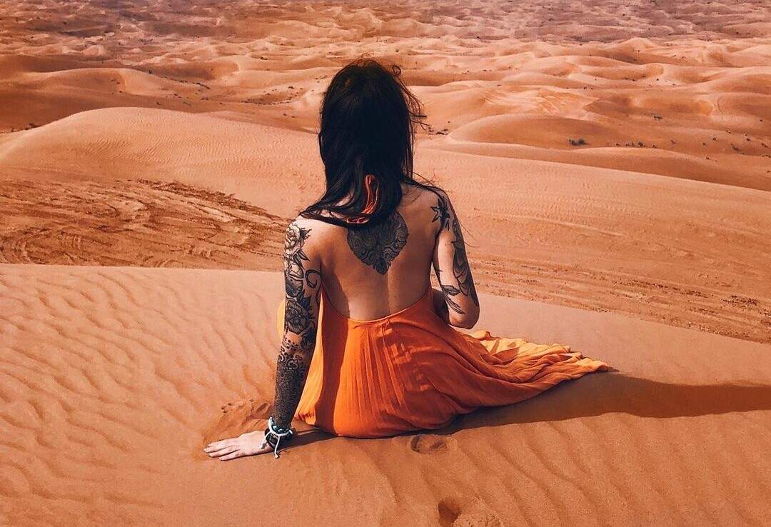 A lone figure with an orange dress and tatted sleeves and back sits on the sloping sand, staring out over the huge desert below.