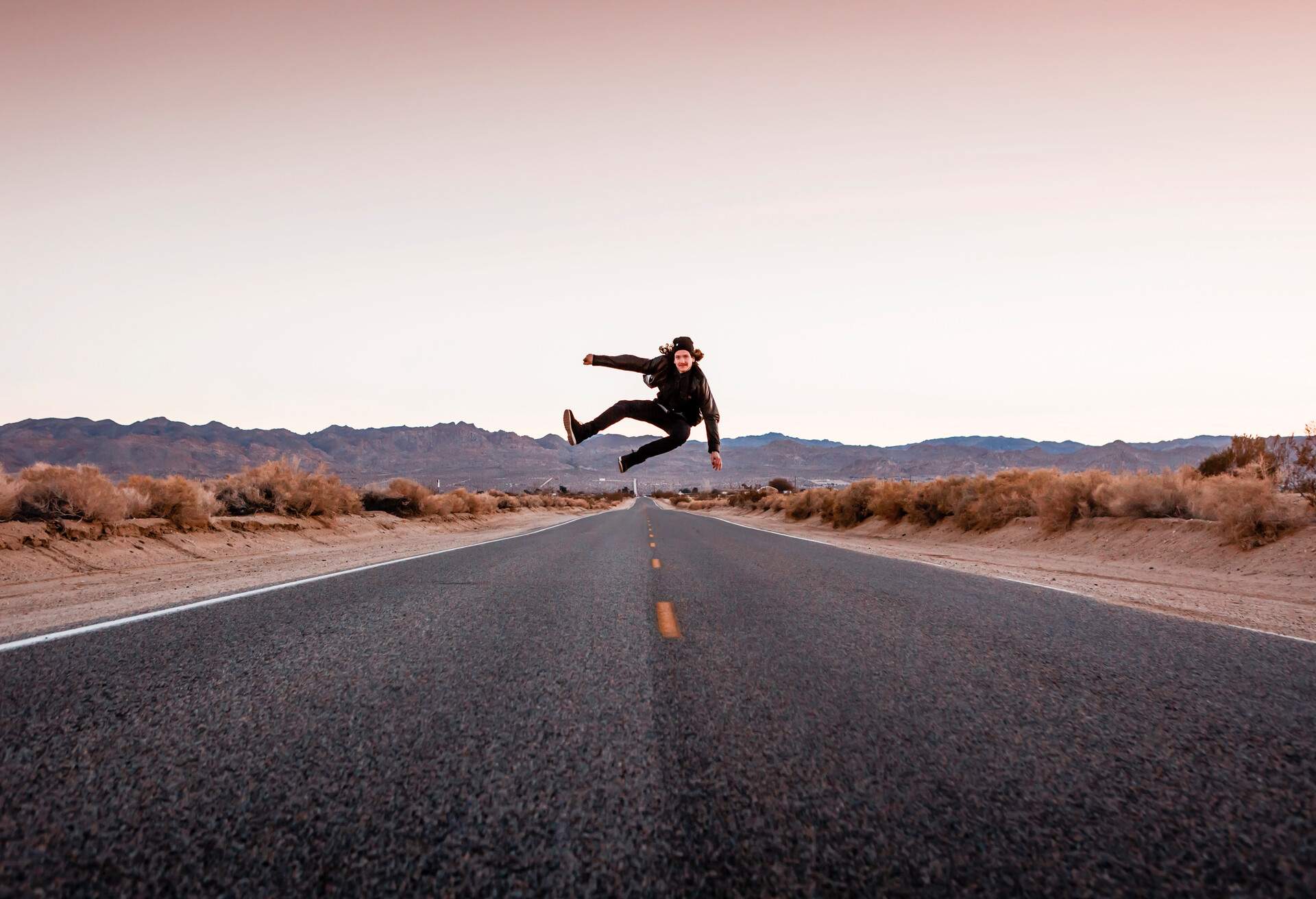 A man in a black jacket and denim pants jumps in the middle of an empty road.