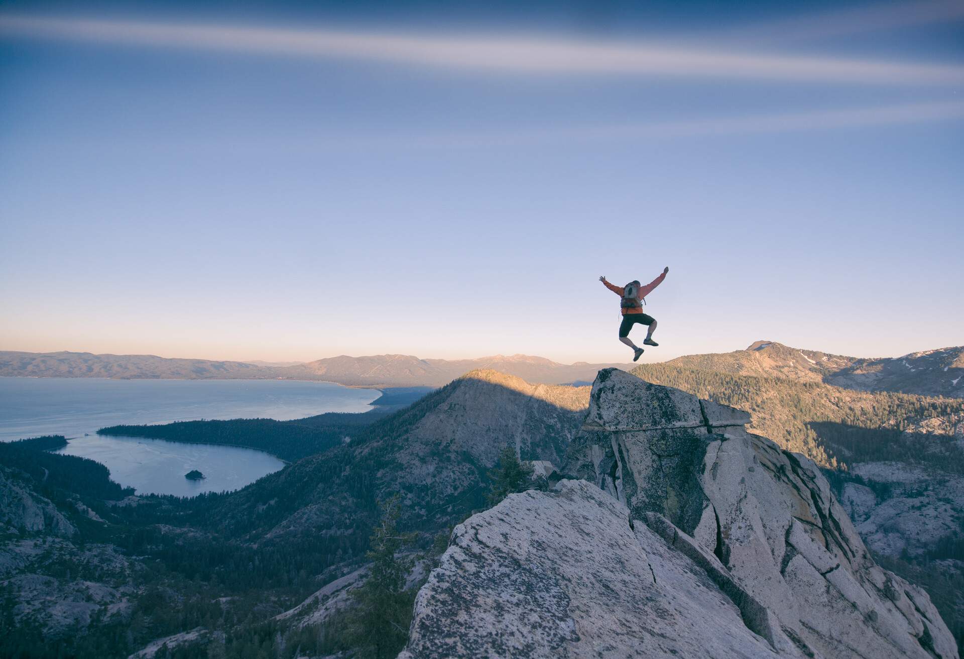 A man jumps on top of a rocky summit with overlooking views of a lake surrounded by a mountainous terrain.