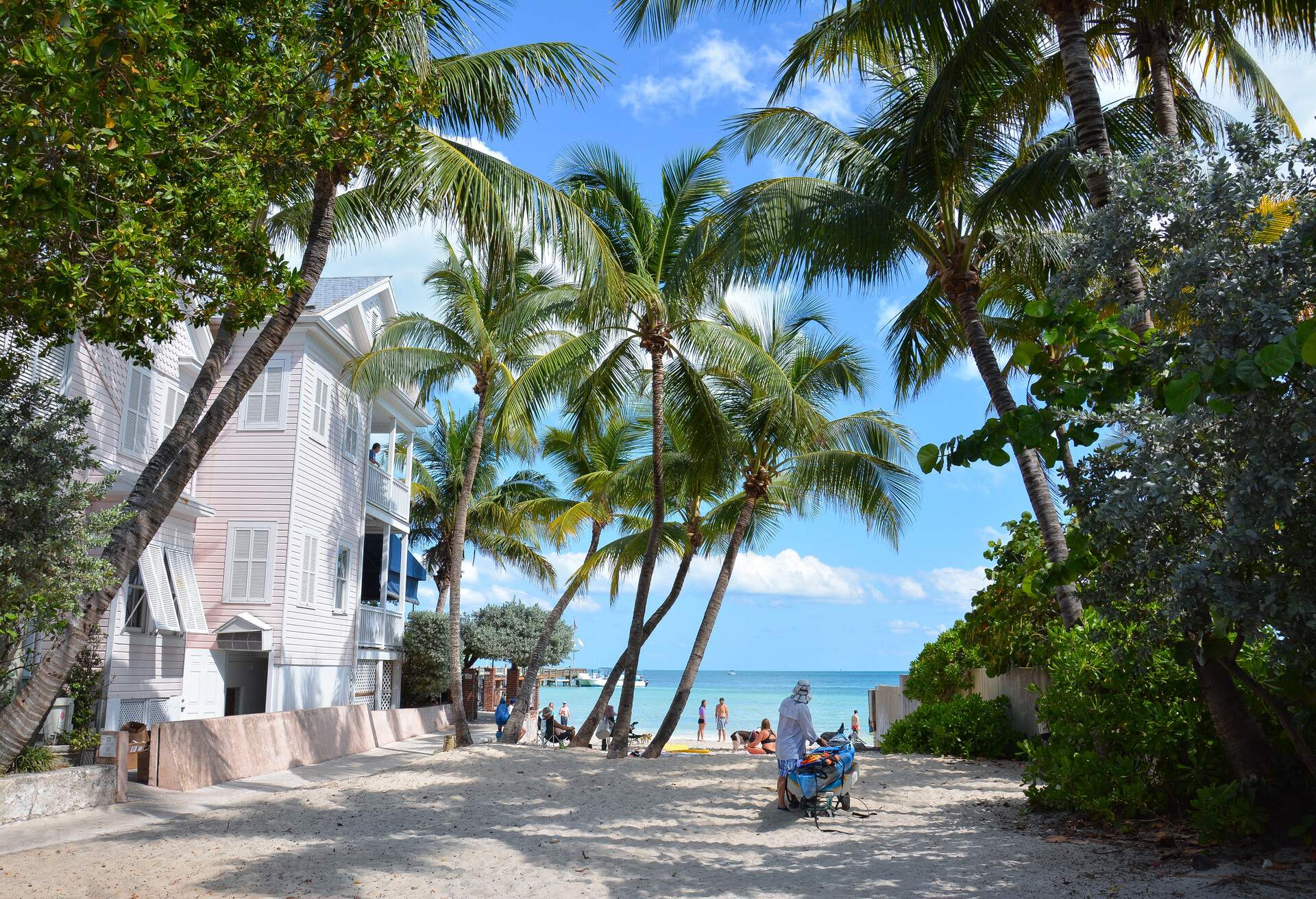 People relax on a beach under a grove of palm trees beside a white beach house.