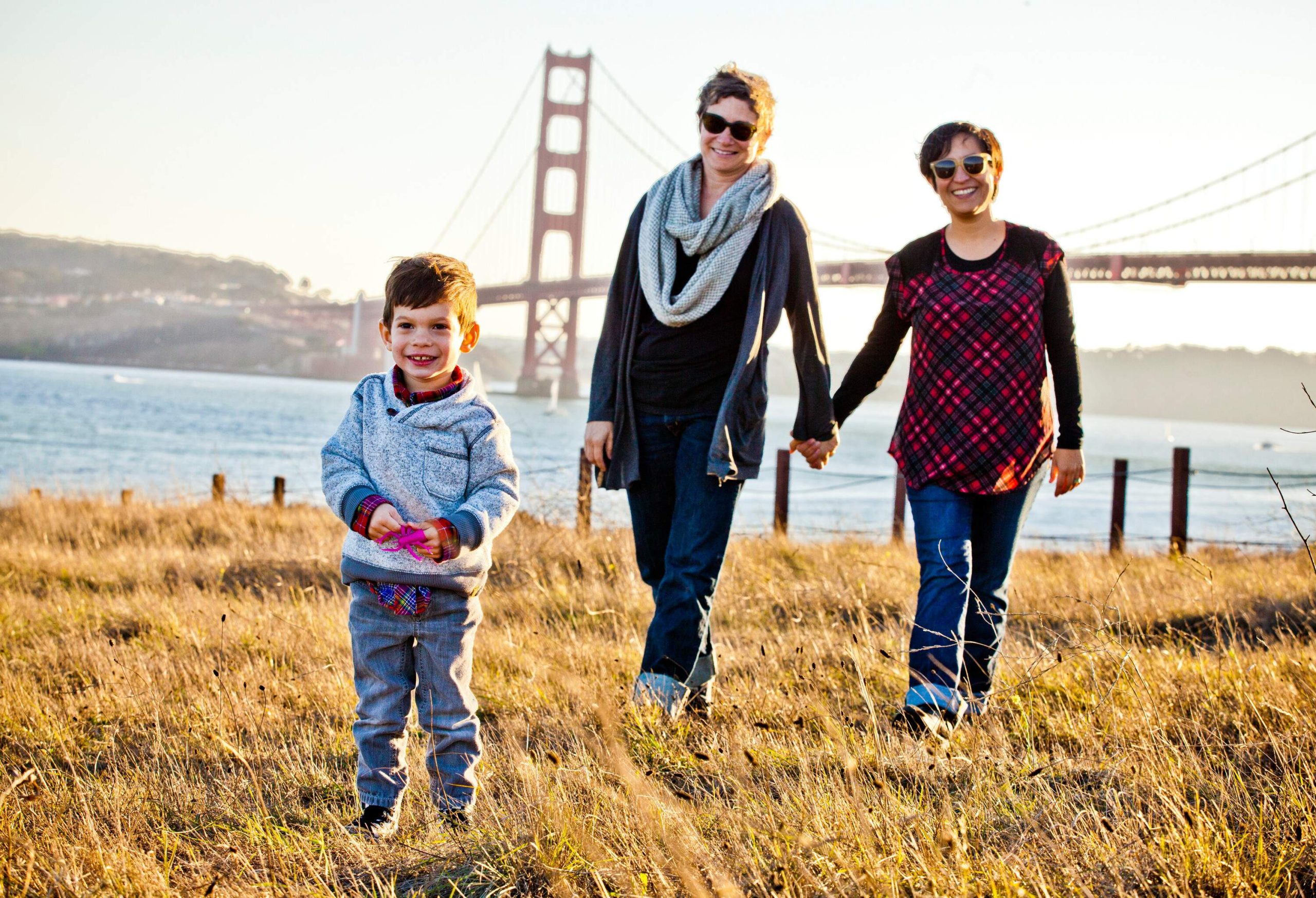 A family of three standing on a grassland with the San Francisco bridge in the distance.
