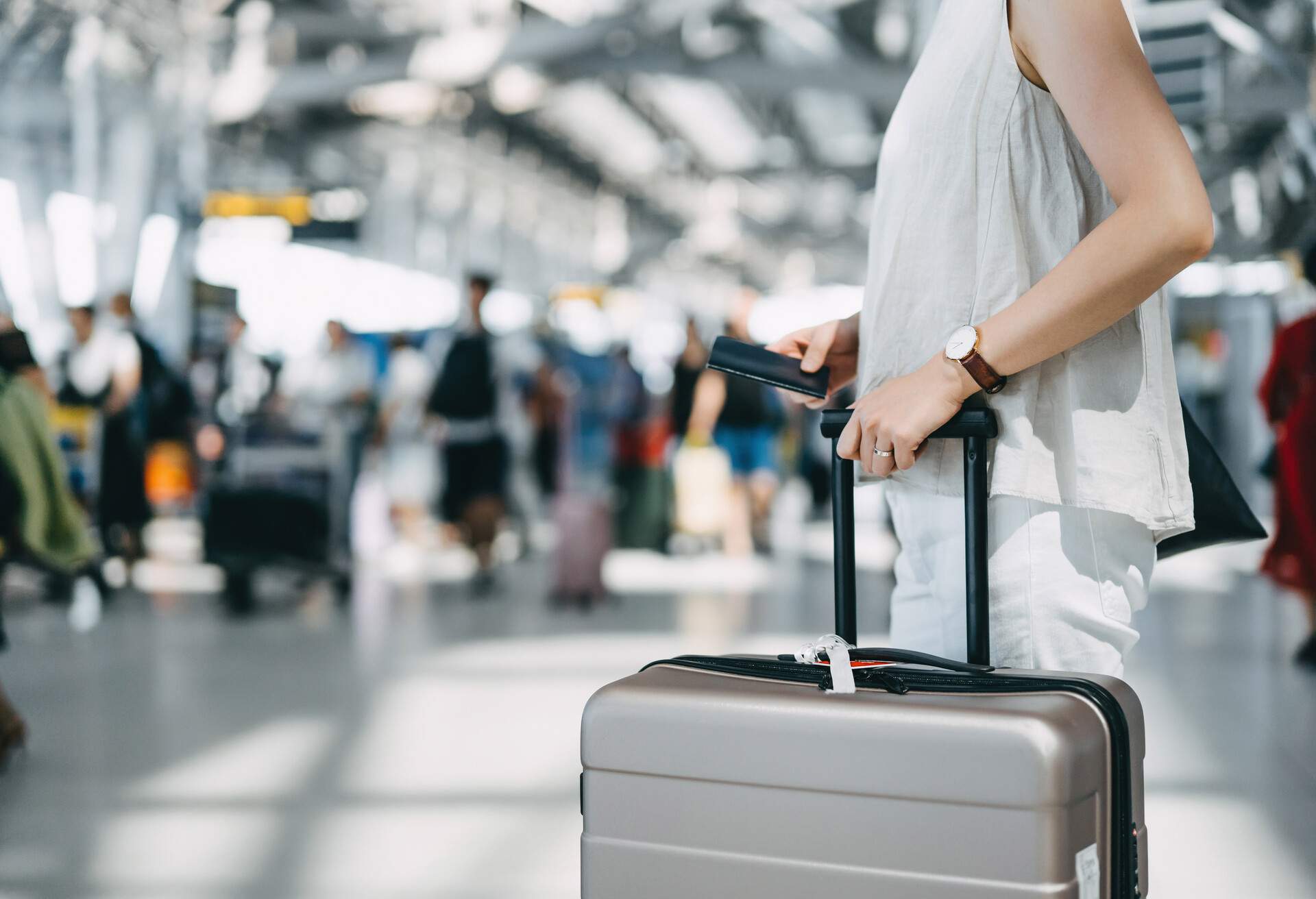 In the bustling international airport hall, a young woman confidently walks with her passport in hand, pulling her suitcase beside her, as the busy surroundings blur in the background.