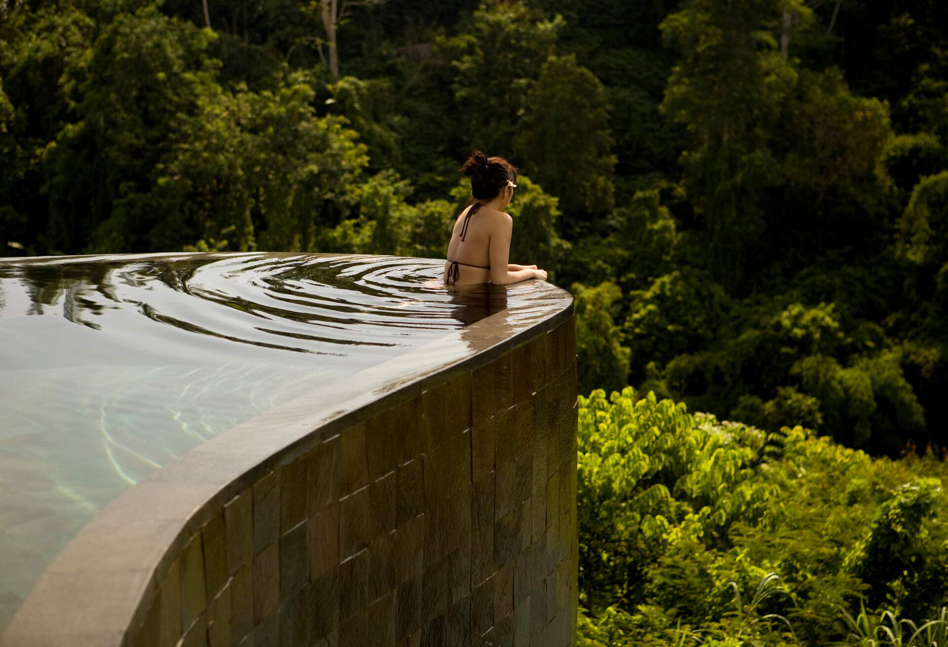 A person in swimwear relax at the edge of a pool and looked out into a jungle in the distance.
