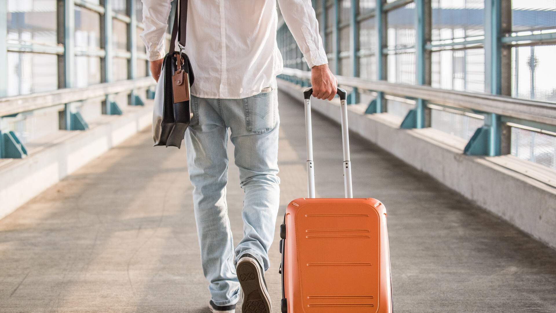 A man wearing casual attire, carrying a backpack and dragging a suitcase, walks along a walkway leading to a railway station.