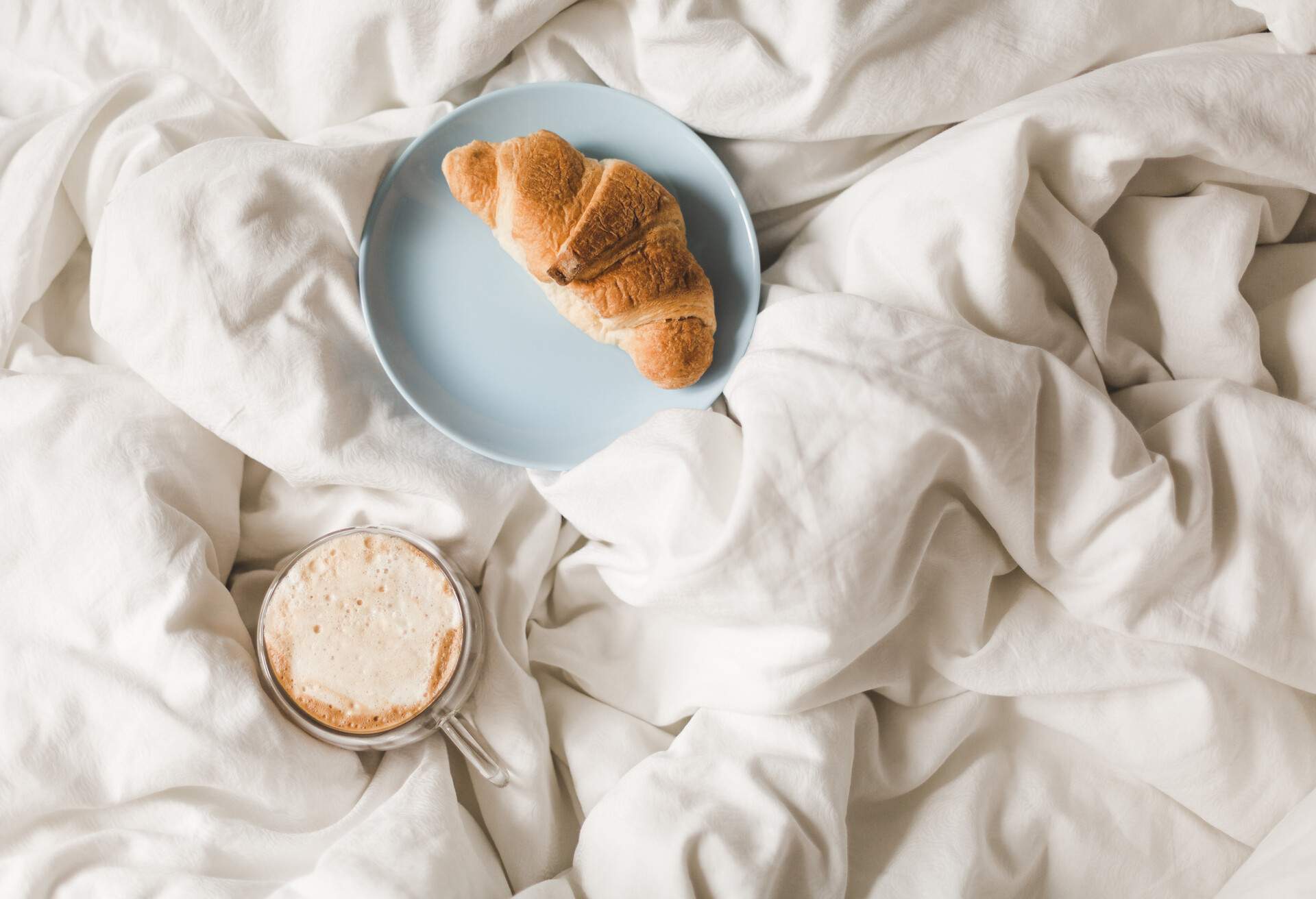 A croissant bread on a blue plate and a cup of coffee served on the white bed.