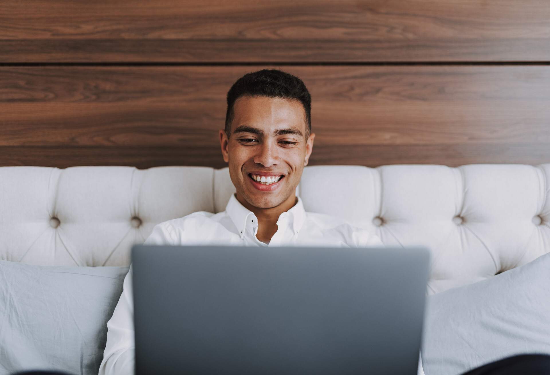 Focus on smiling handsome man relaxing on bedroom. He is holding laptop and looking at screen while networking.
