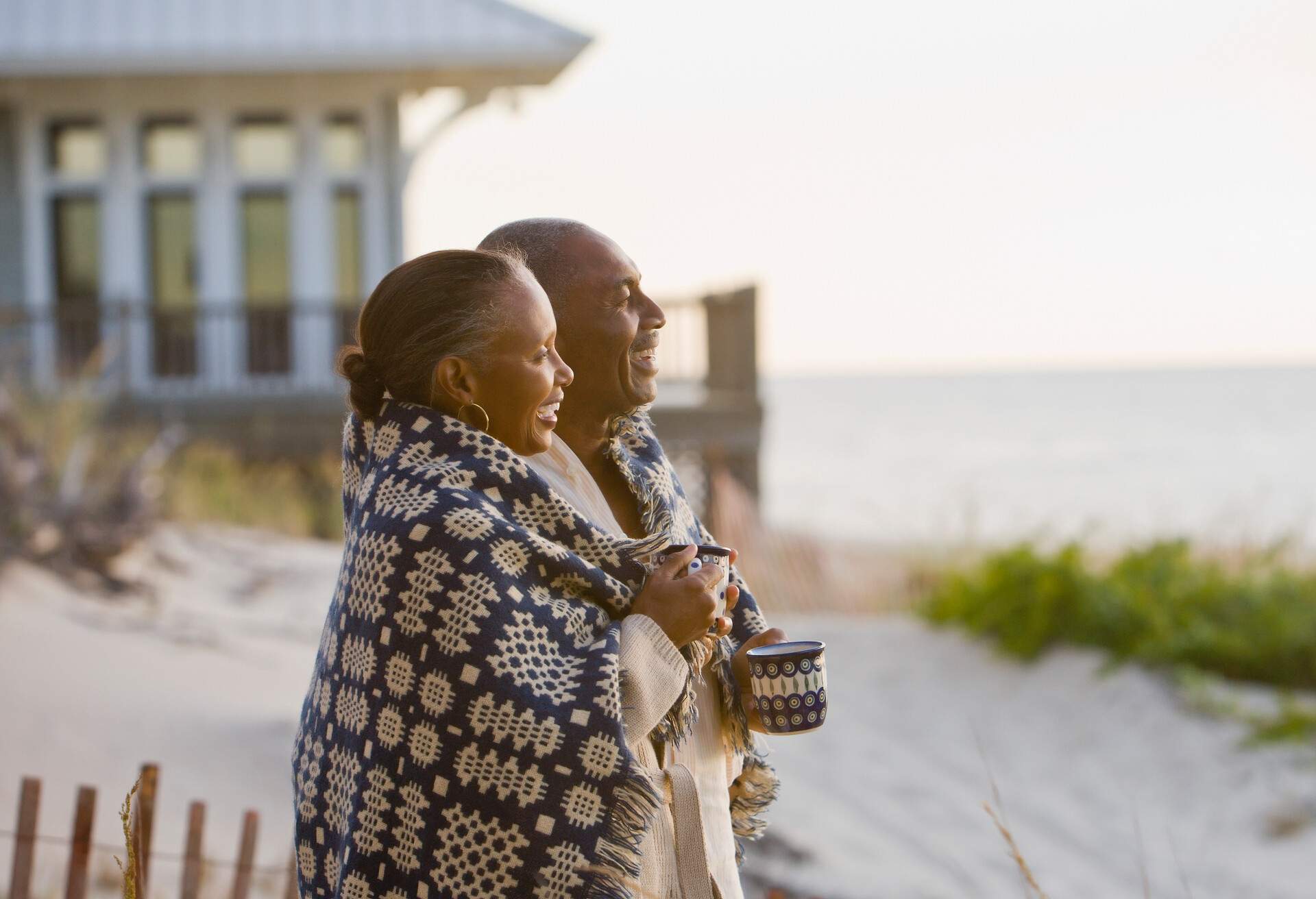 An elderly couple wrapped themselves with a knitted shawl as they smile on a beach.