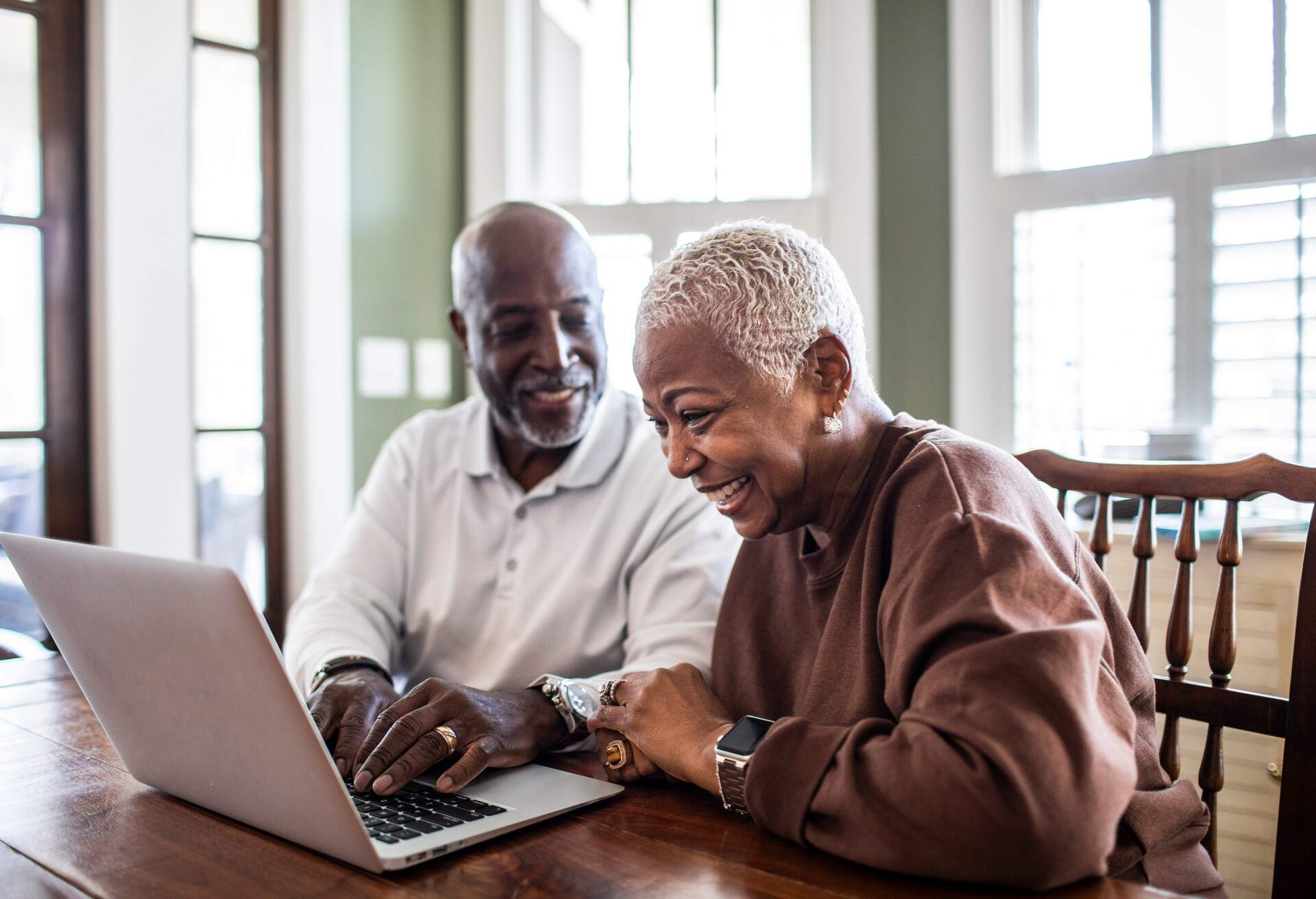 A happy elderly couple using a laptop that sits on a wooden table.