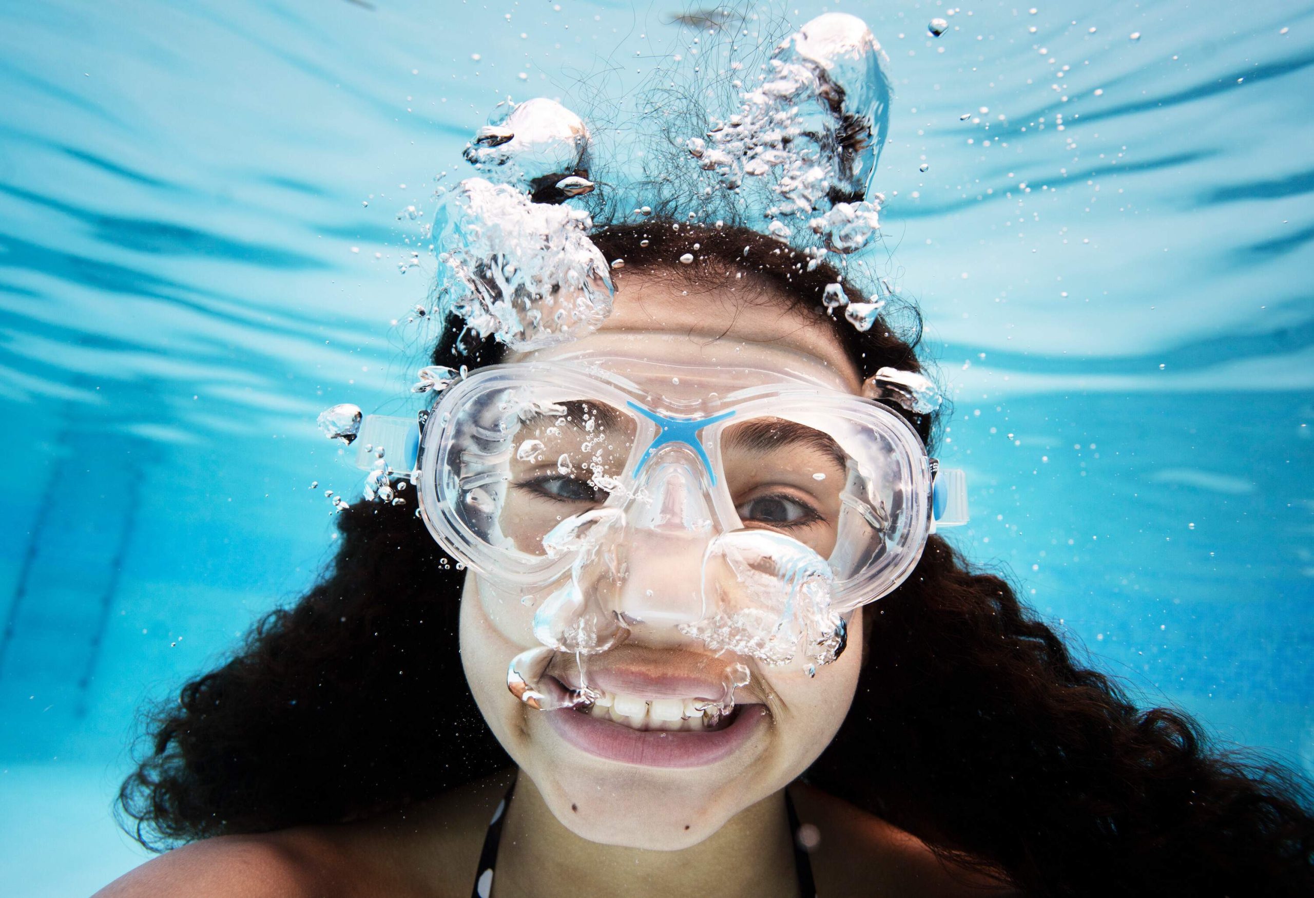 A teen girl smiles at the camera while blowing bubbles underwater.