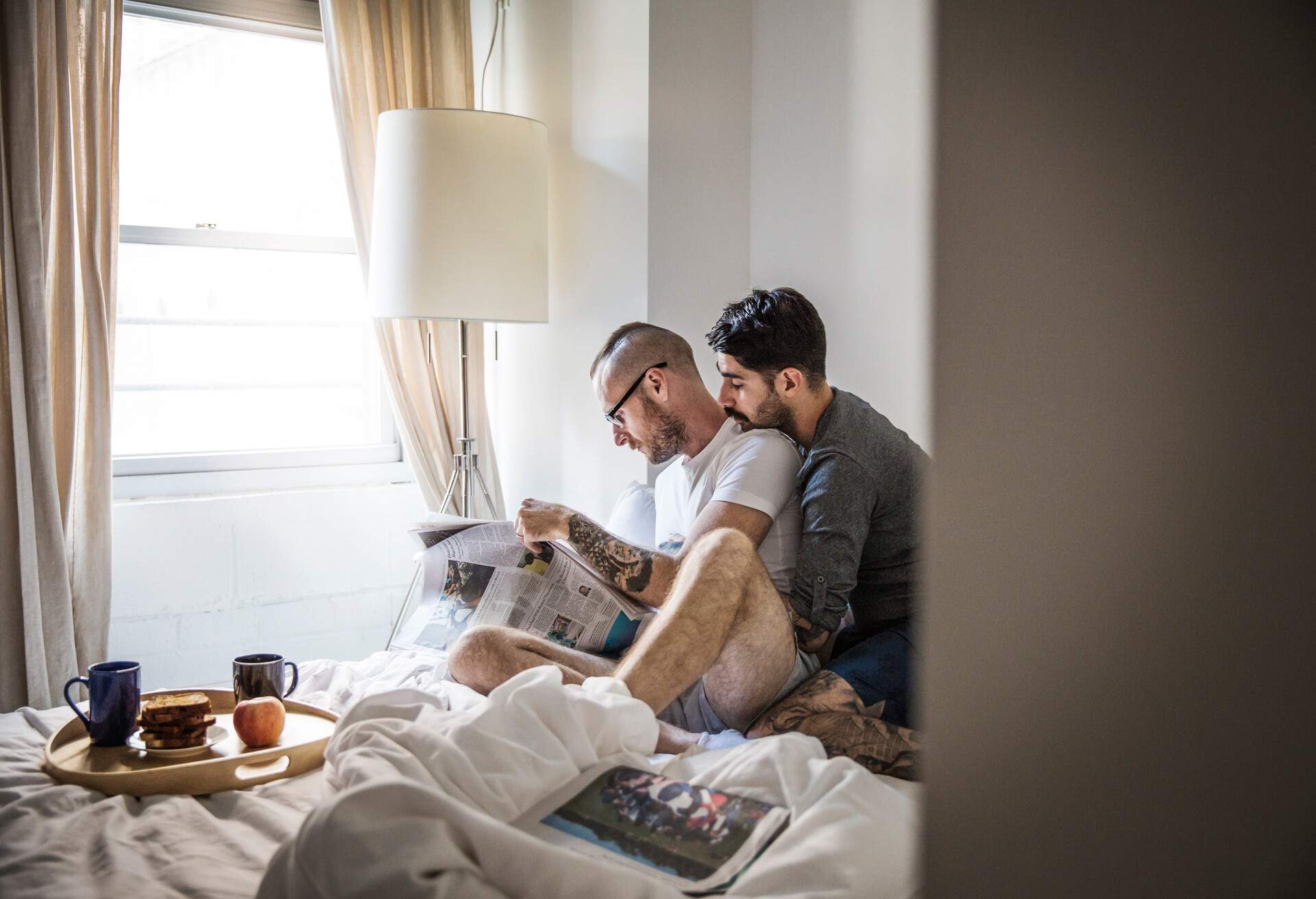 A person hugging his boyfriend from behind while sitting on the bed with breakfast on the tray and reading the newspaper.