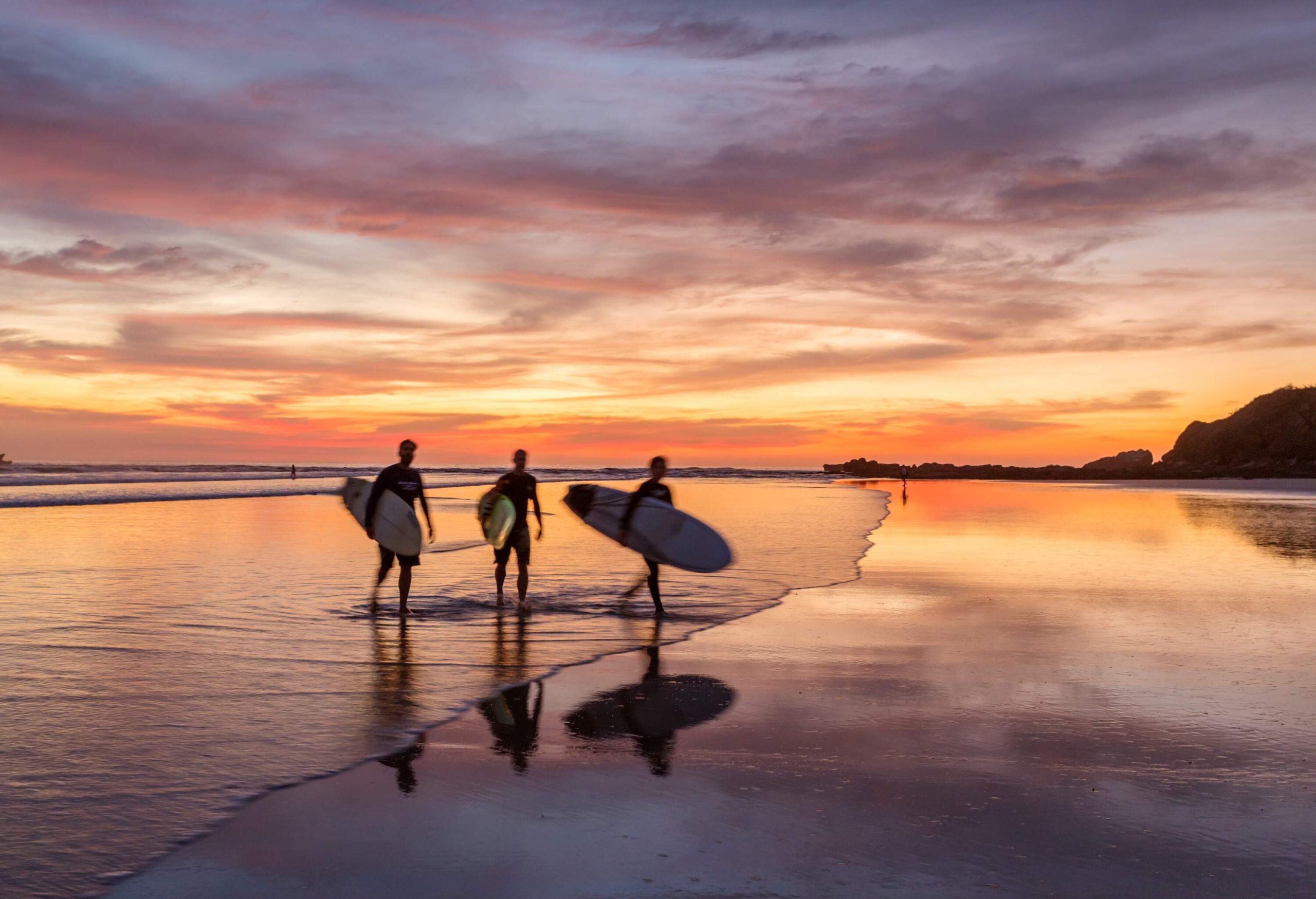 Three male surfers carrying their boards after surfing, walking towards the shore against the dramatic sky.