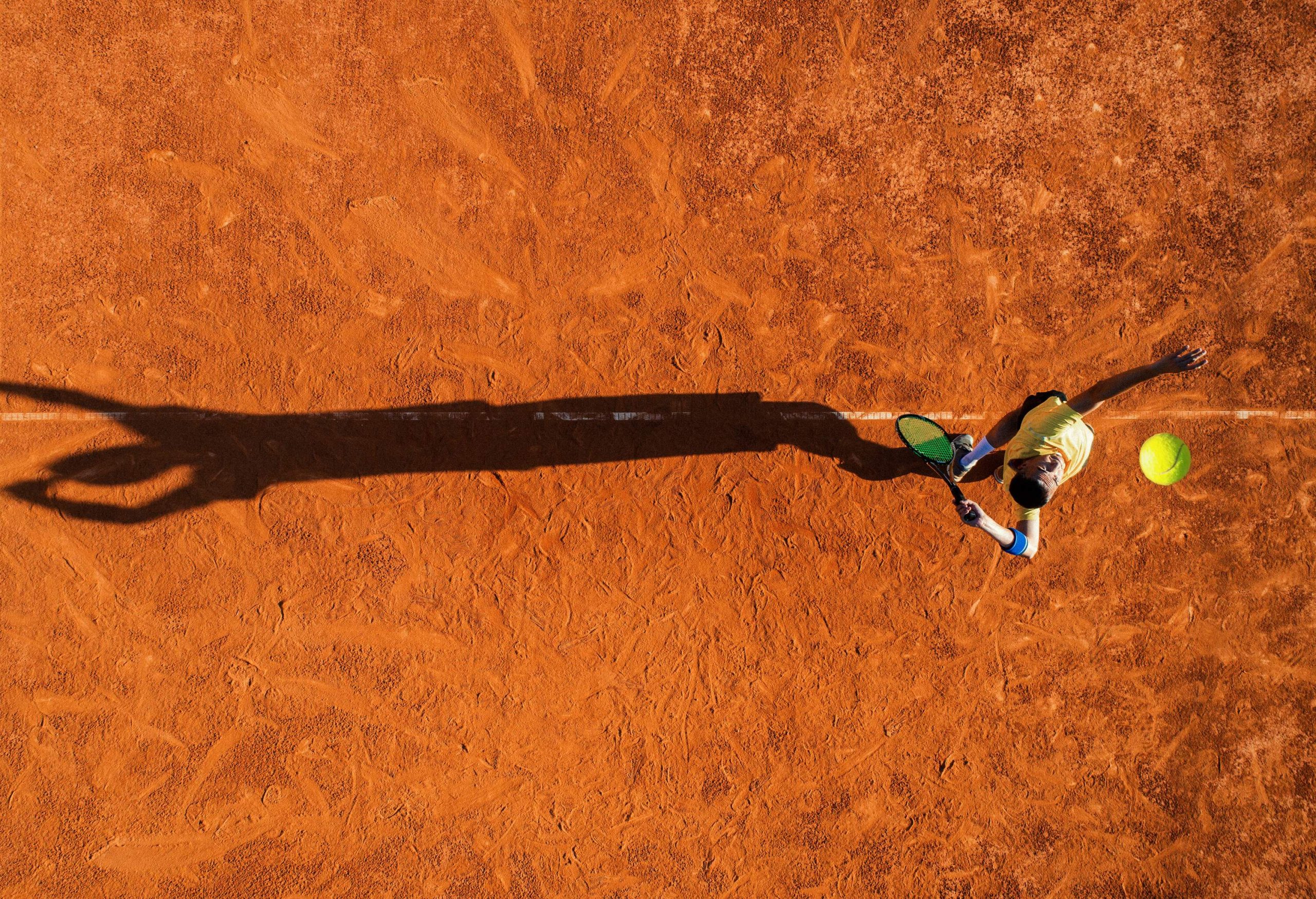 A tennis player serving on a clay court with his shadow cast on the ground.