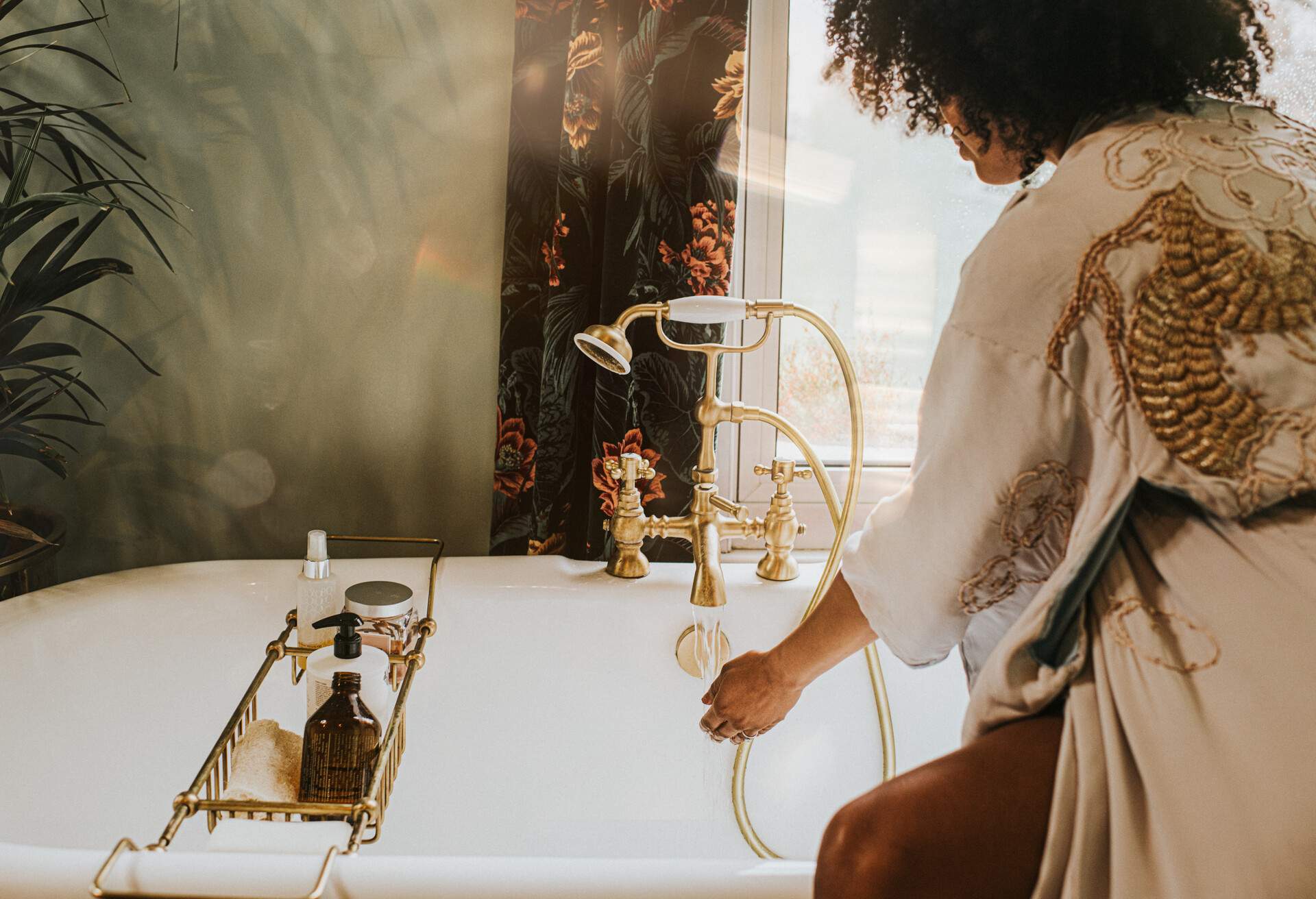 A curly-haired woman in a bathrobe checks the bathtub water's temperature.