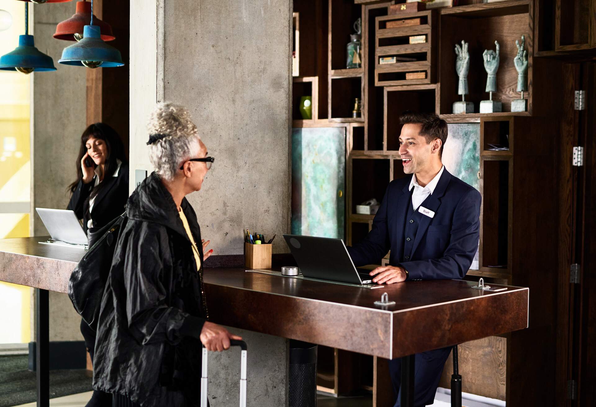A male hotel receptionist in a navy blue suit talks to an elderly woman behind the counter.